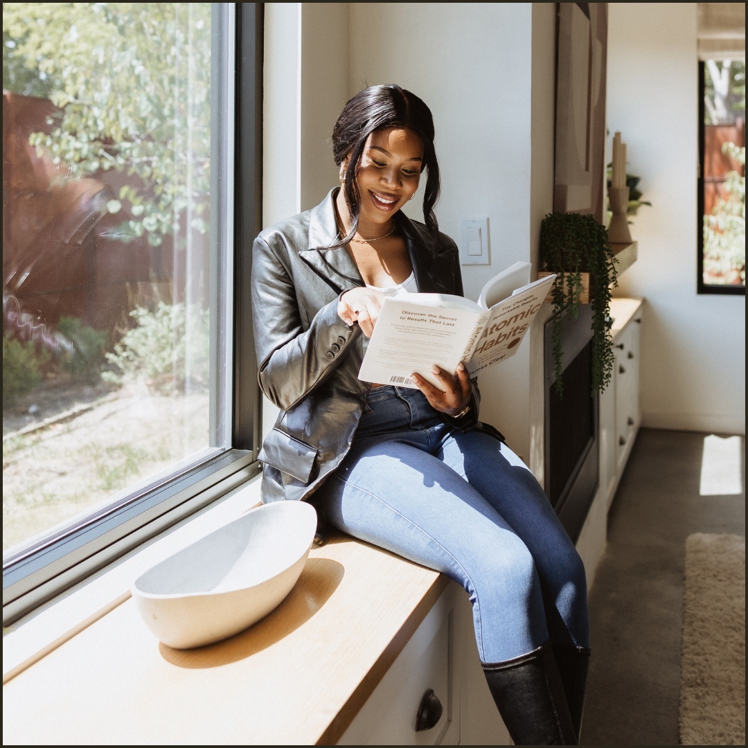 Dheandra Nicolette sitting on a kitchen window ledge, reading a book and smiling. She wears a black leather jacket, white top, and blue jeans. Sunlight streams through the window, illuminating the space.