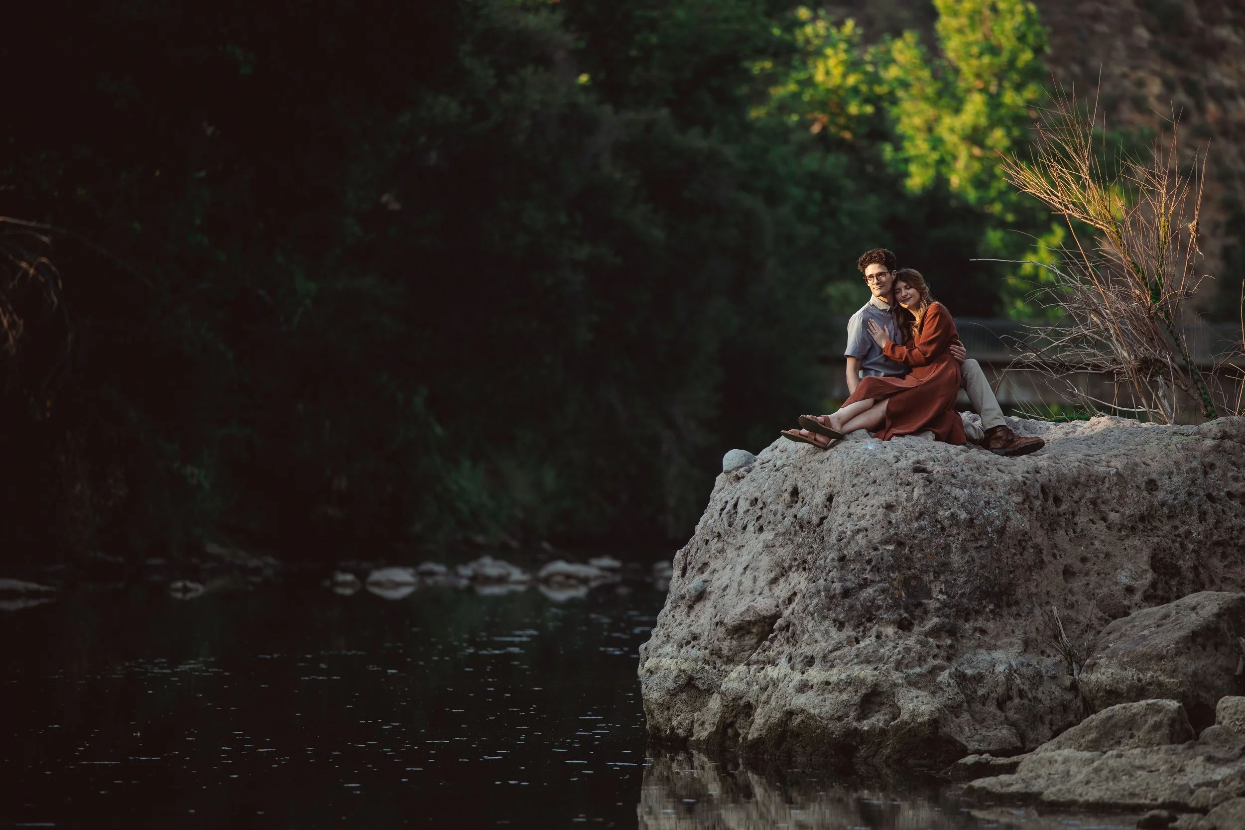 A young couple sitting on a large rock by a river, embracing and enjoying a peaceful moment surrounded by trees.