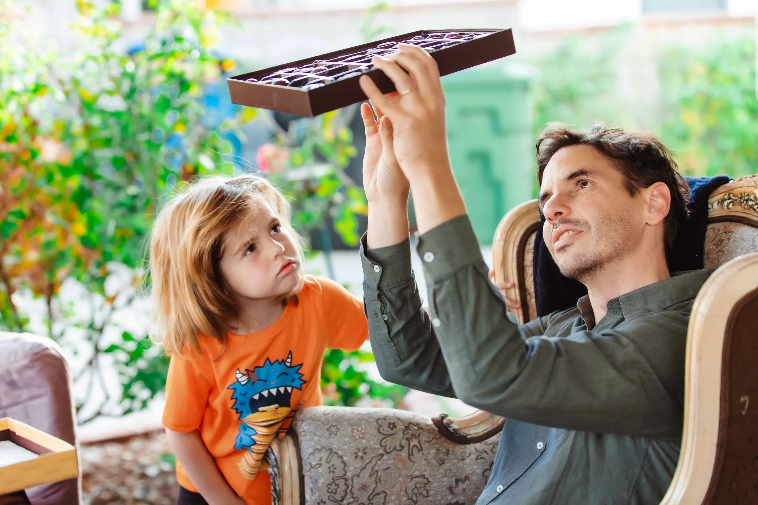 A young girl with an orange shirt watching a man hold a chocolate box, seated outdoors with greenery in the background.