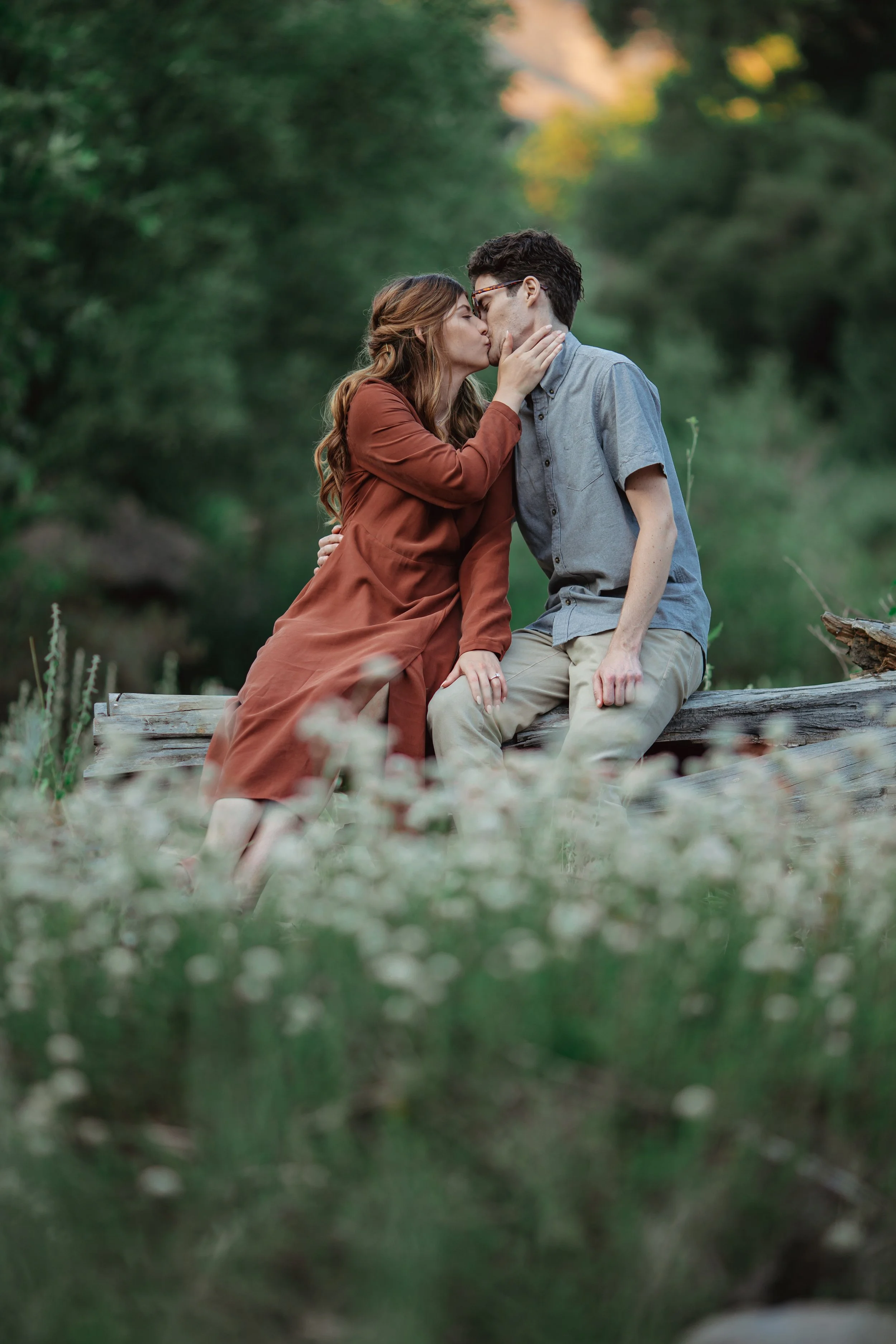 A couple sitting on a log in a forest, sharing a kiss