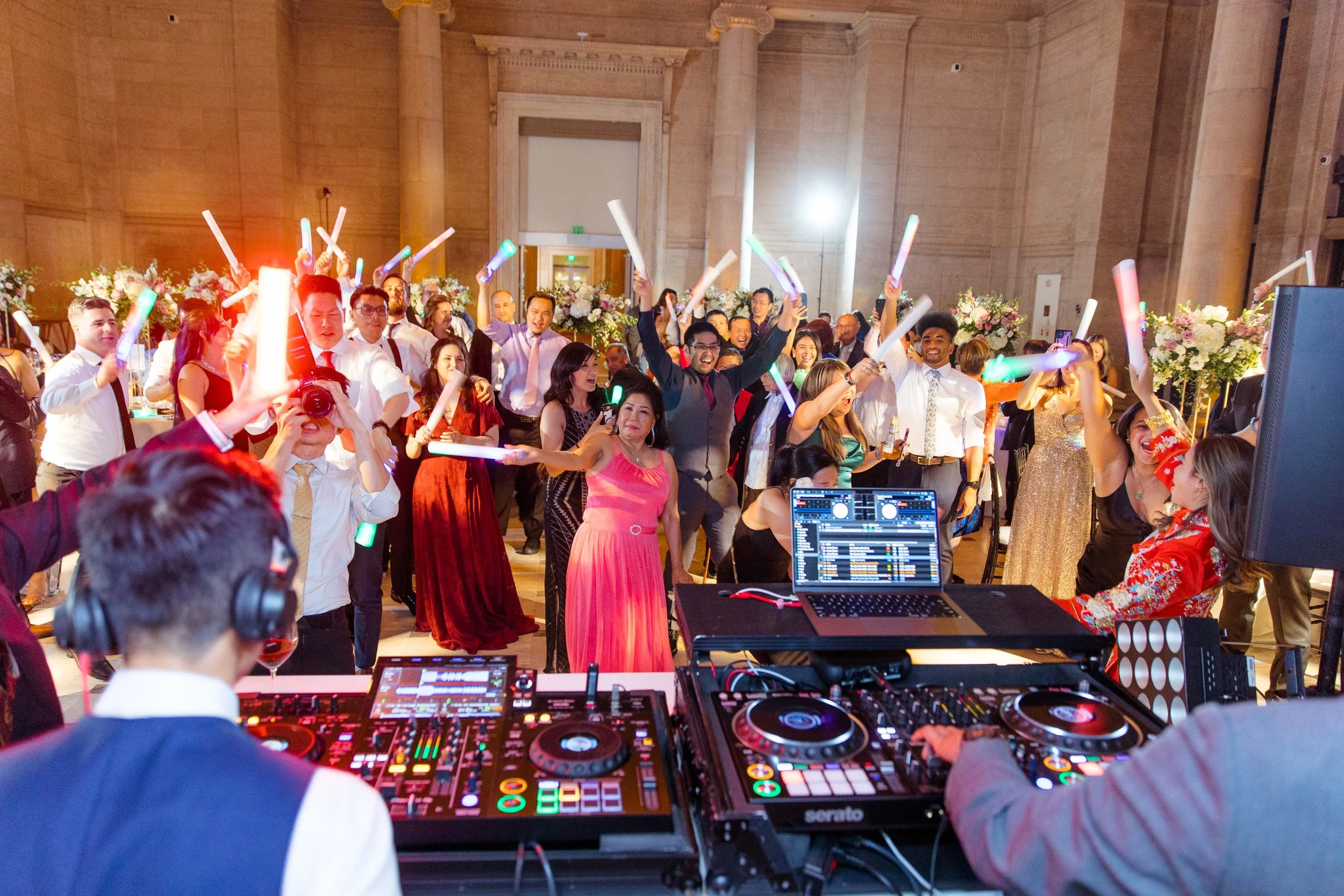 People dancing and celebrating at a wedding reception with DJ equipment in the foreground and floral decorations in the background.