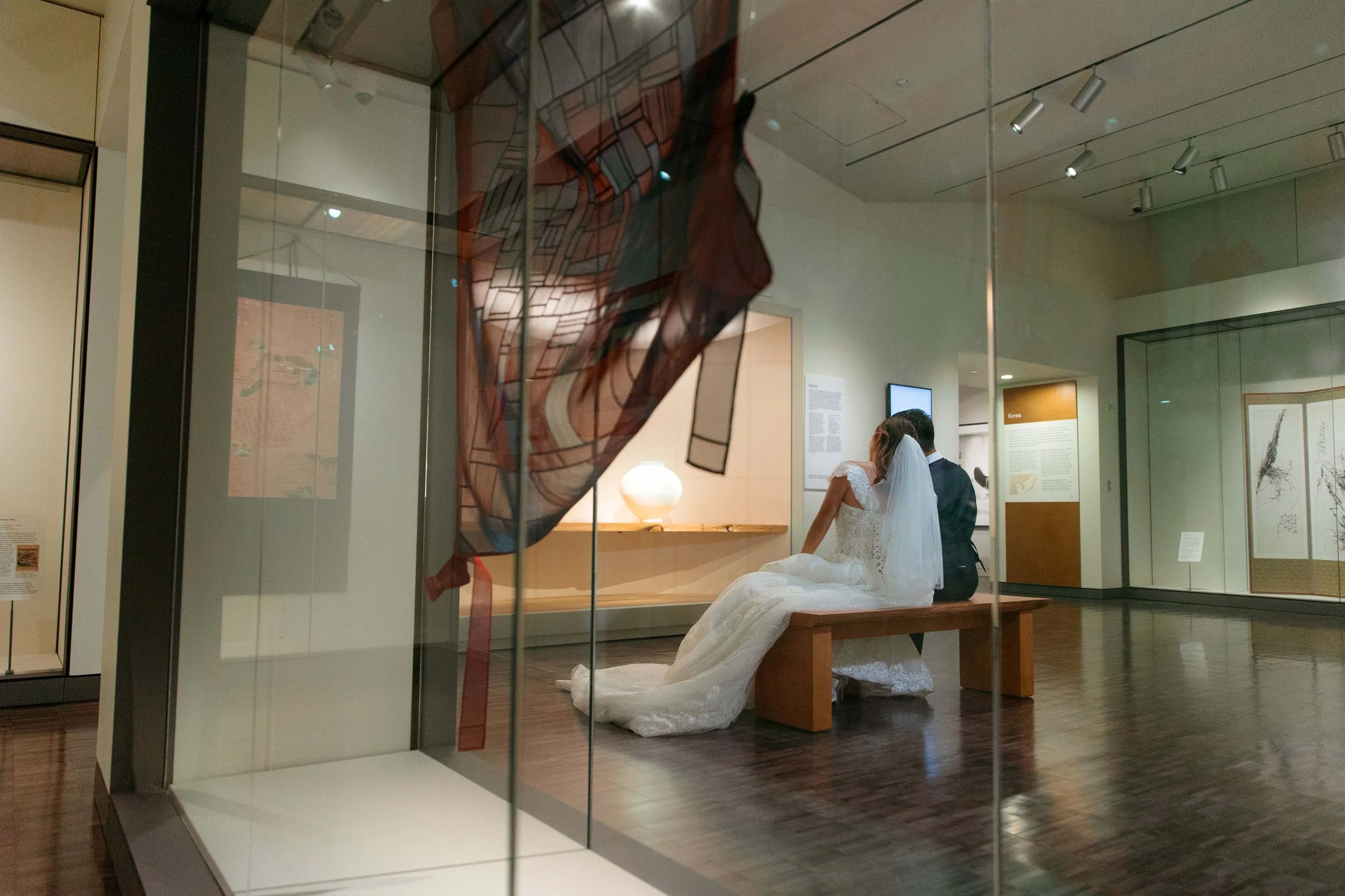 A bride and groom sitting on a wooden bench in a museum, with the bride dressed in a white wedding gown and veil, and the groom in a dark suit, viewed through a glass display case with artwork inside.