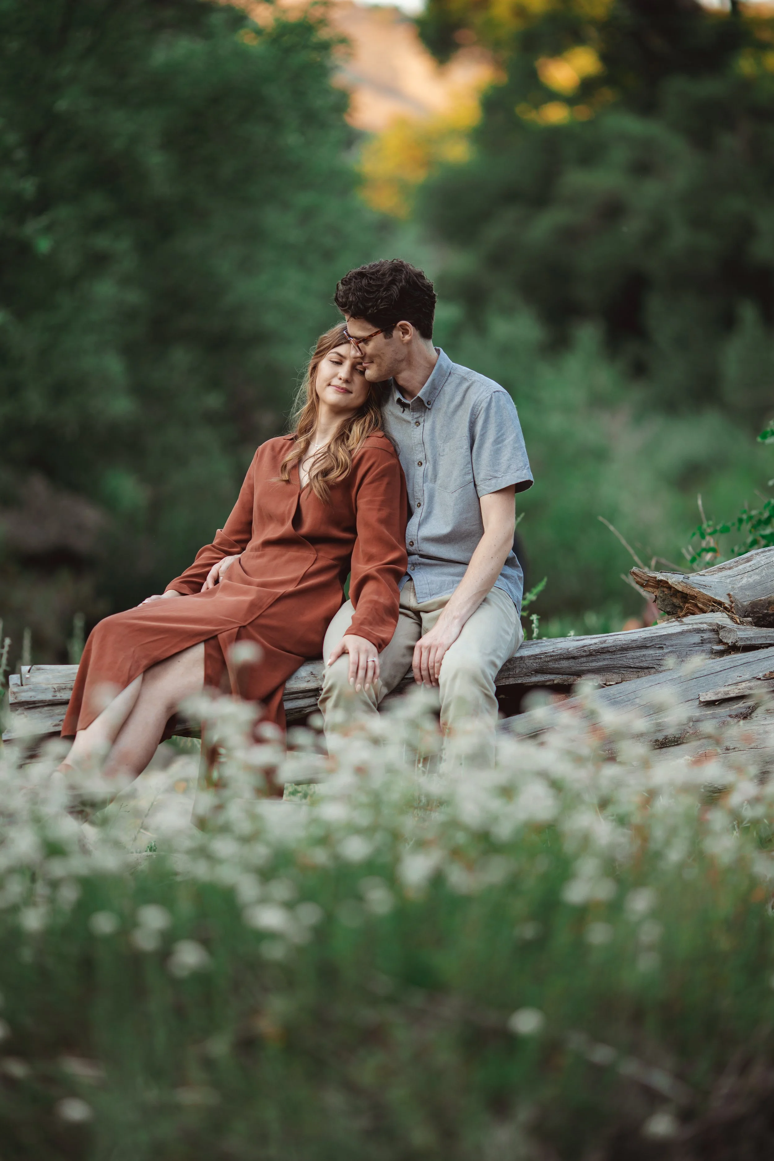 A young couple sits close together on a fallen log in a forest setting, with trees and greenery in the background. The woman is leaning her head against the man's, with both appearing peaceful and content.