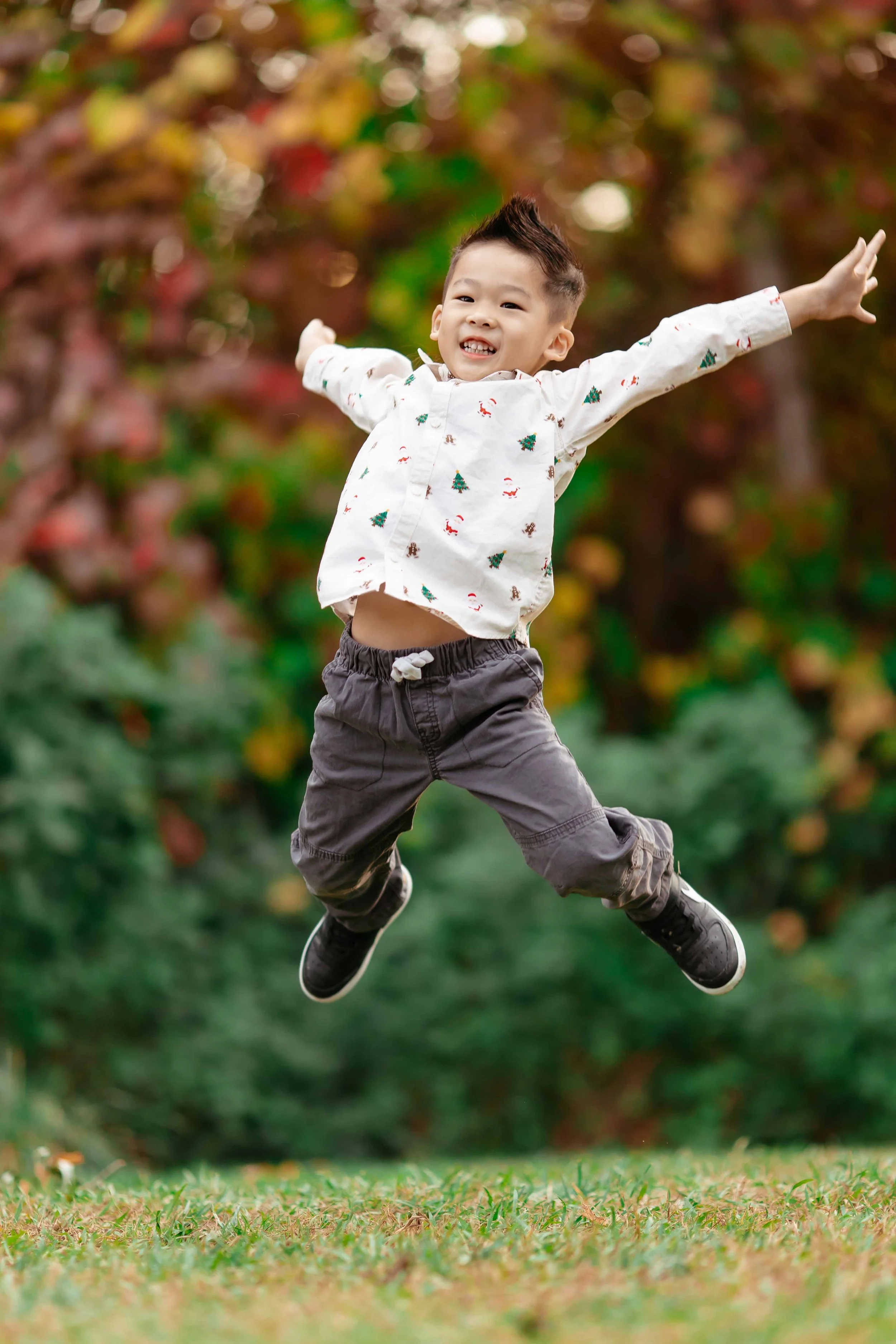 A young boy is jumping with arms wide open on a grassy field, with a colorful fall foliage background.