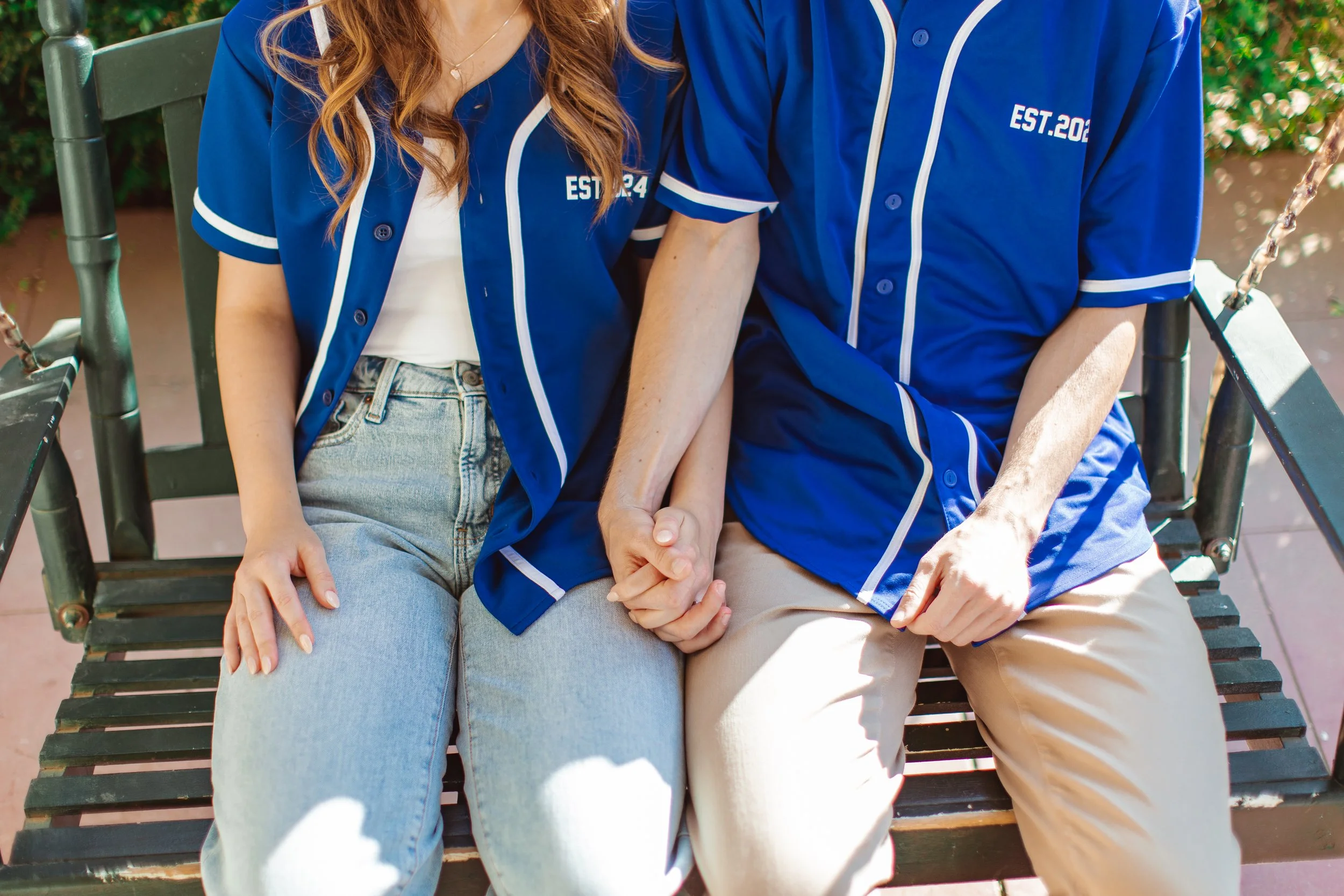 A couple sitting on a bench holding hands, both wearing matching blue baseball jerseys.