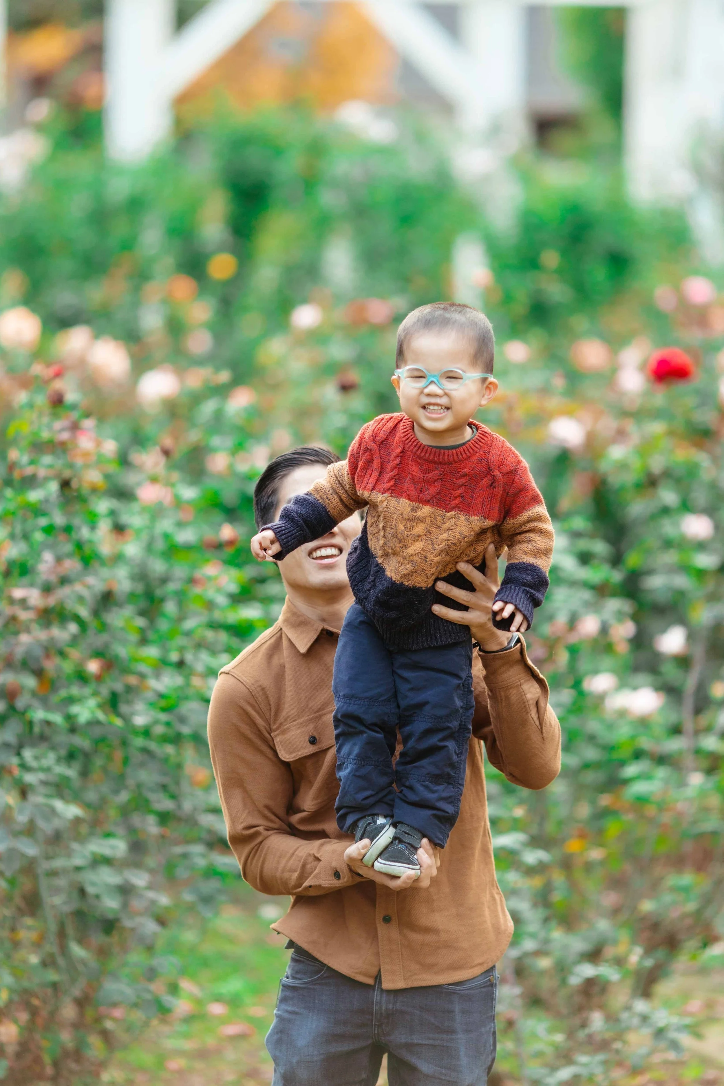 A man holding a smiling child with glasses in a rose garden.