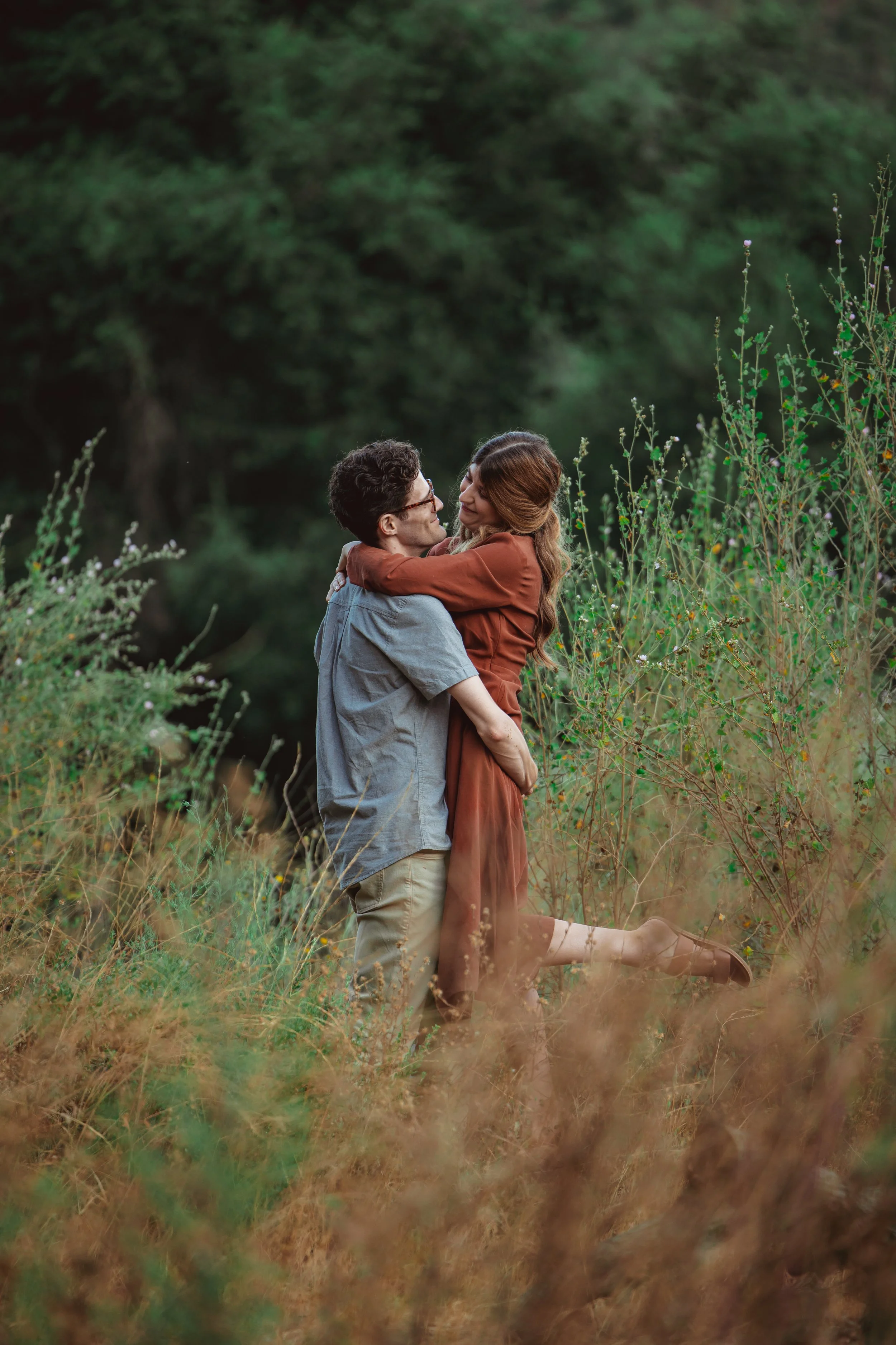 A couple embracing in a green, outdoor setting. The woman is lifted off the ground, wearing a rust-colored dress, and the man is in a casual shirt and tan pants. They are surrounded by tall grasses and bushes with a forested background.