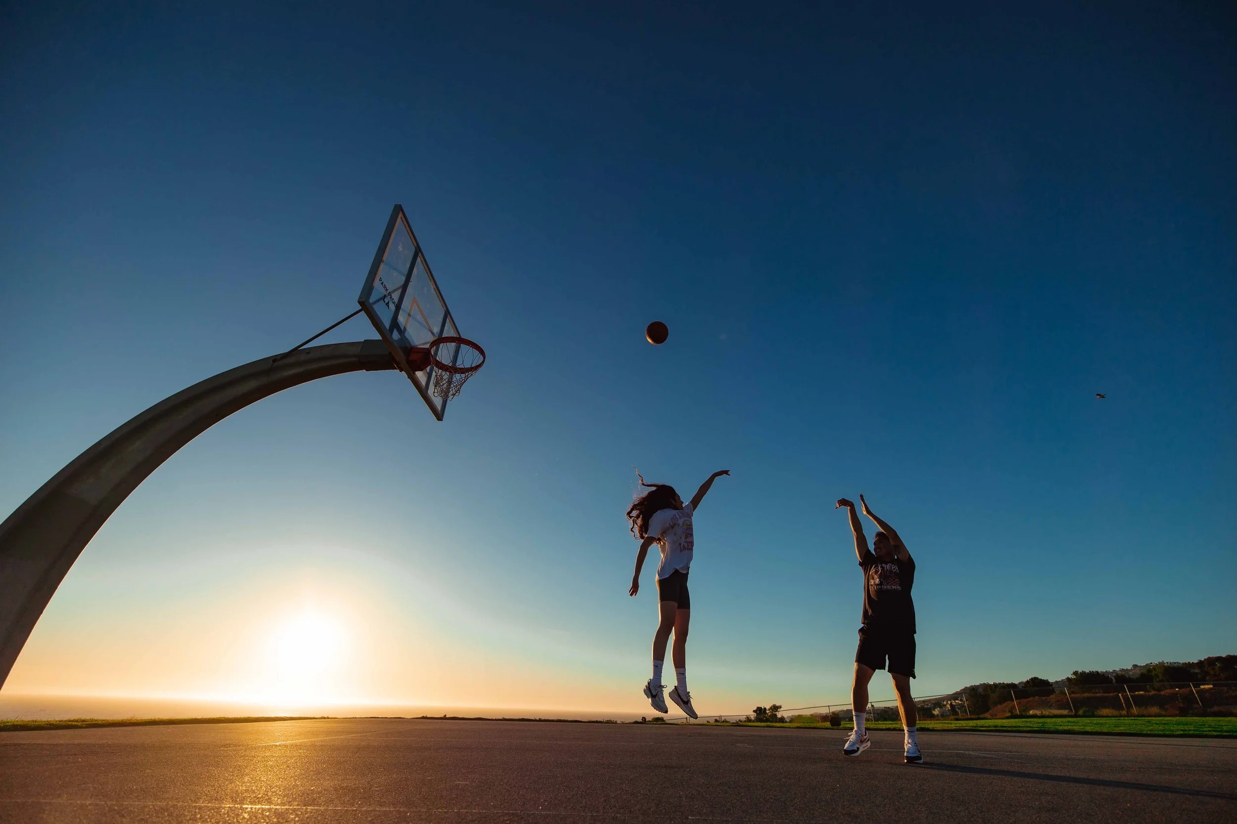 Two children playing basketball on an outdoor court at sunset, with one child jumping to shoot while the other reaches to block, and the basketball in mid-air near the hoop.