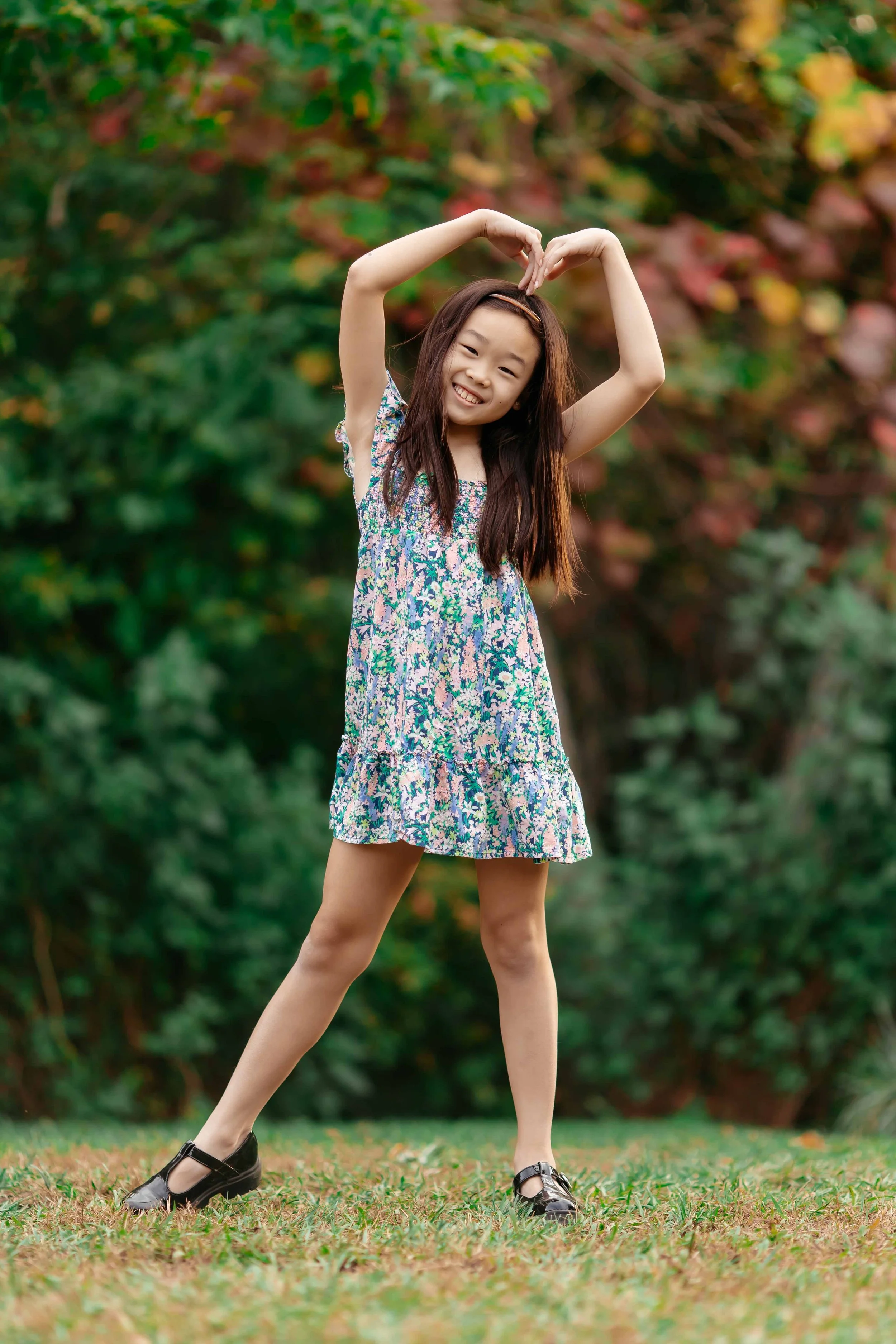 A young girl standing outdoors on grass during fall, forming a heart shape over her head with her arms, smiling at the camera, wearing a floral dress and black Mary Jane shoes, with autumn-colored trees in the background.