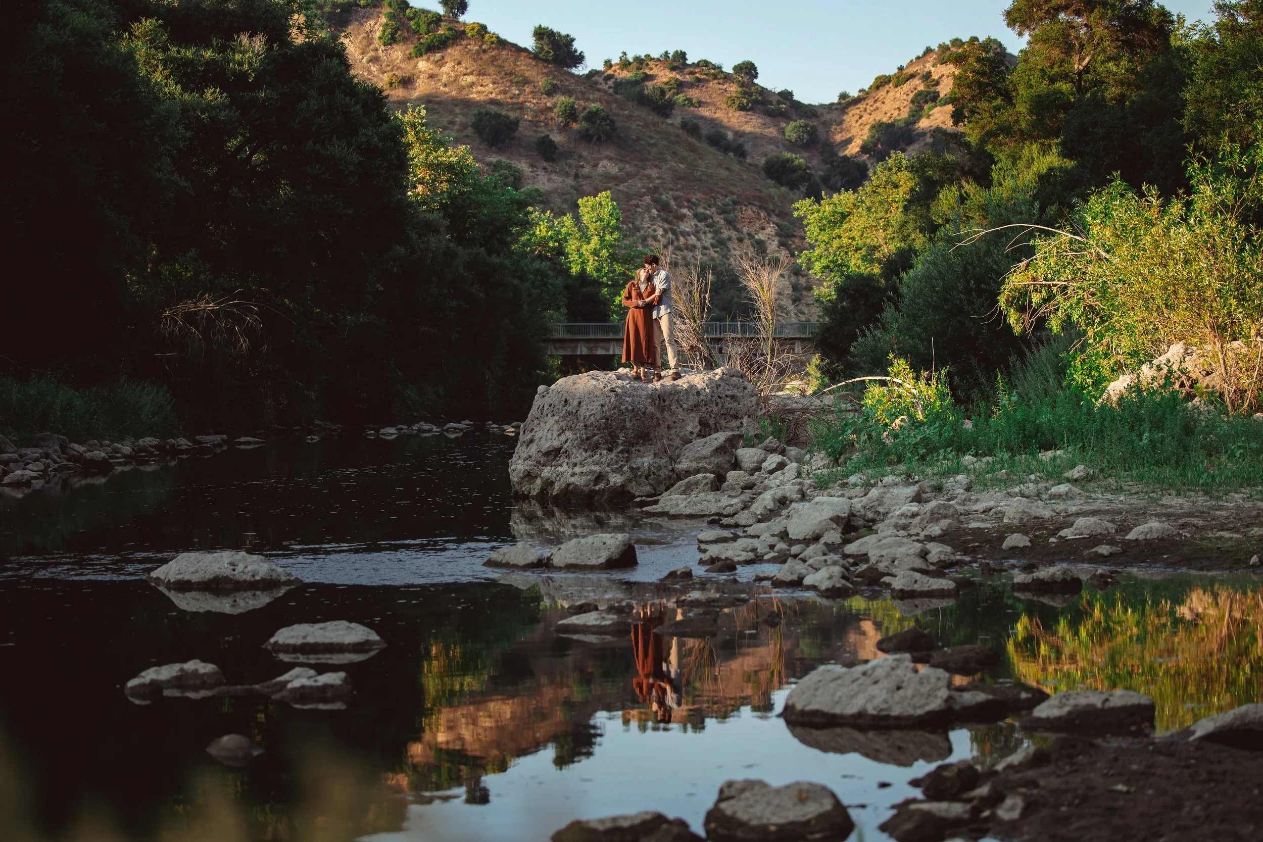A couple standing on a large rock in the middle of a river, surrounded by lush green trees and hills, with a bridge in the background, during late afternoon or early evening.