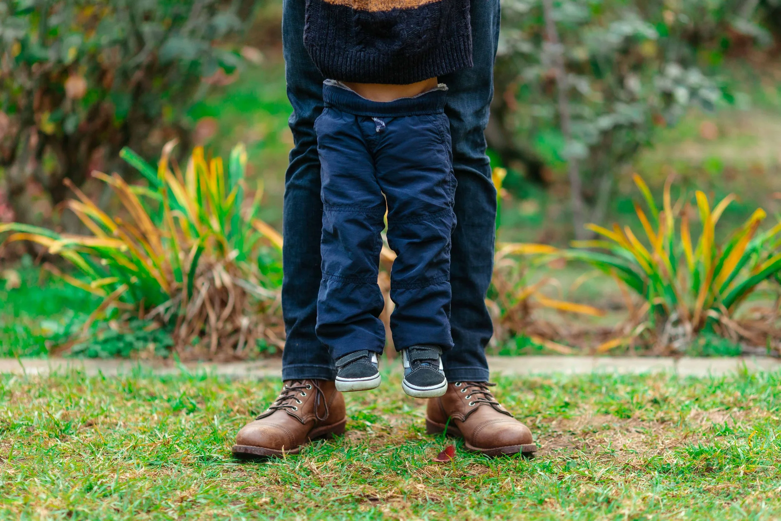 A person standing outdoors with a child hanging upside down from their legs. The person's feet are shown wearing brown boots, and the child is wearing dark pants and sneakers, with the child's head and upper body out of the frame. The background incl