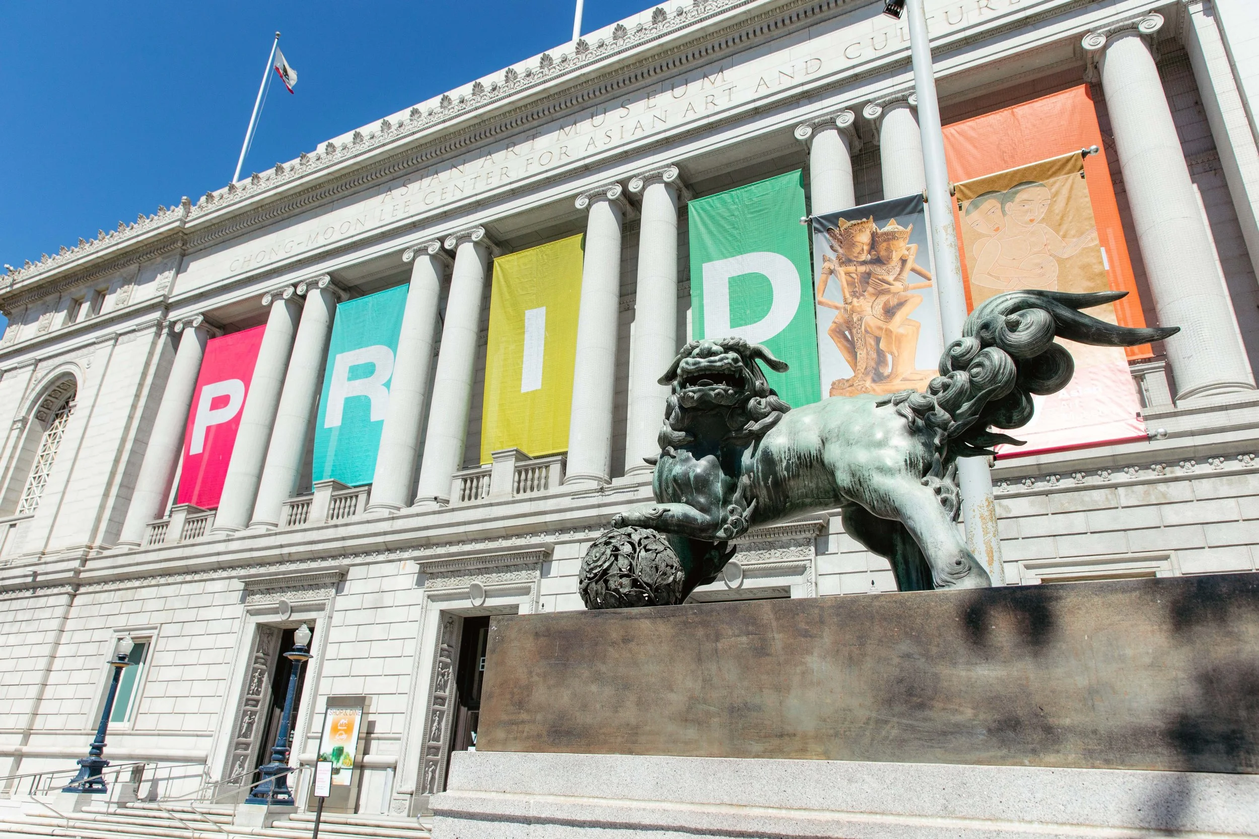 Exterior of the Asian Art Museum in San Francisco with large colorful banners that spell "PRIDE" and a bronze mythical creature statue in front.