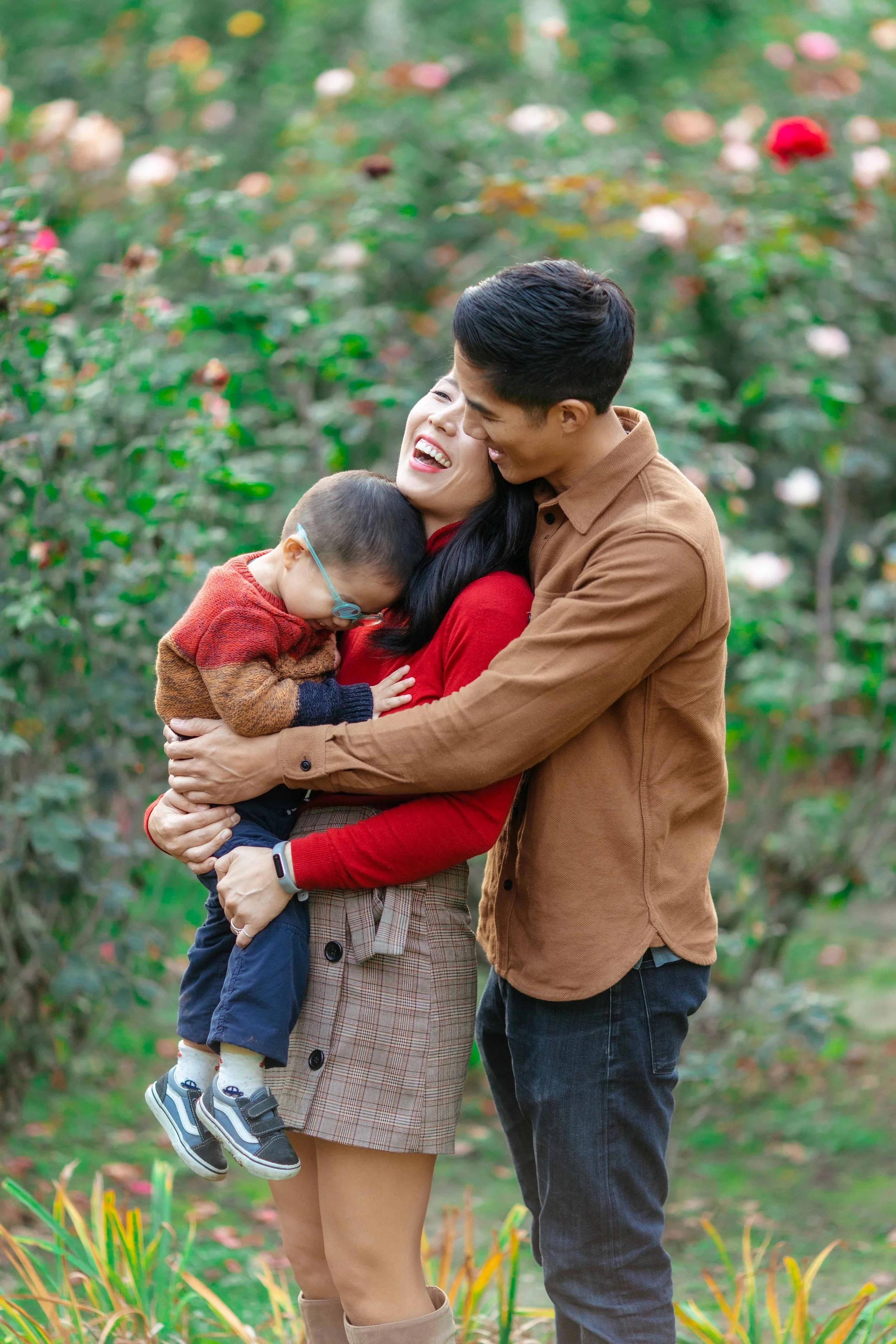 A family of three happily hugging outdoors among blooming flowers and bushes.