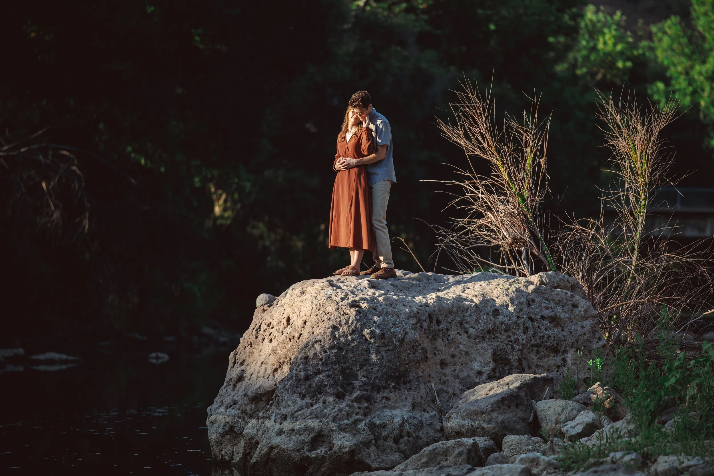 A couple embraces on a large rock by a river in a forested area during sunset, with trees and greenery in the background.