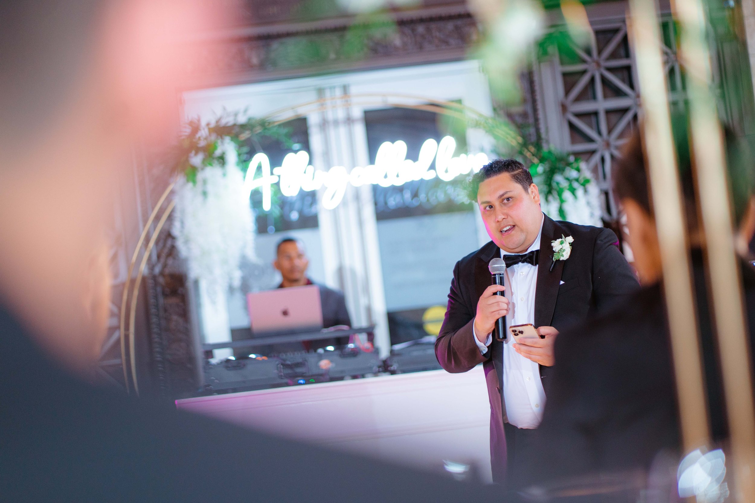 A man in a tuxedo holding a microphone and a phone, speaking at a wedding reception with a DJ in the background, and decorative lighting and plants around.