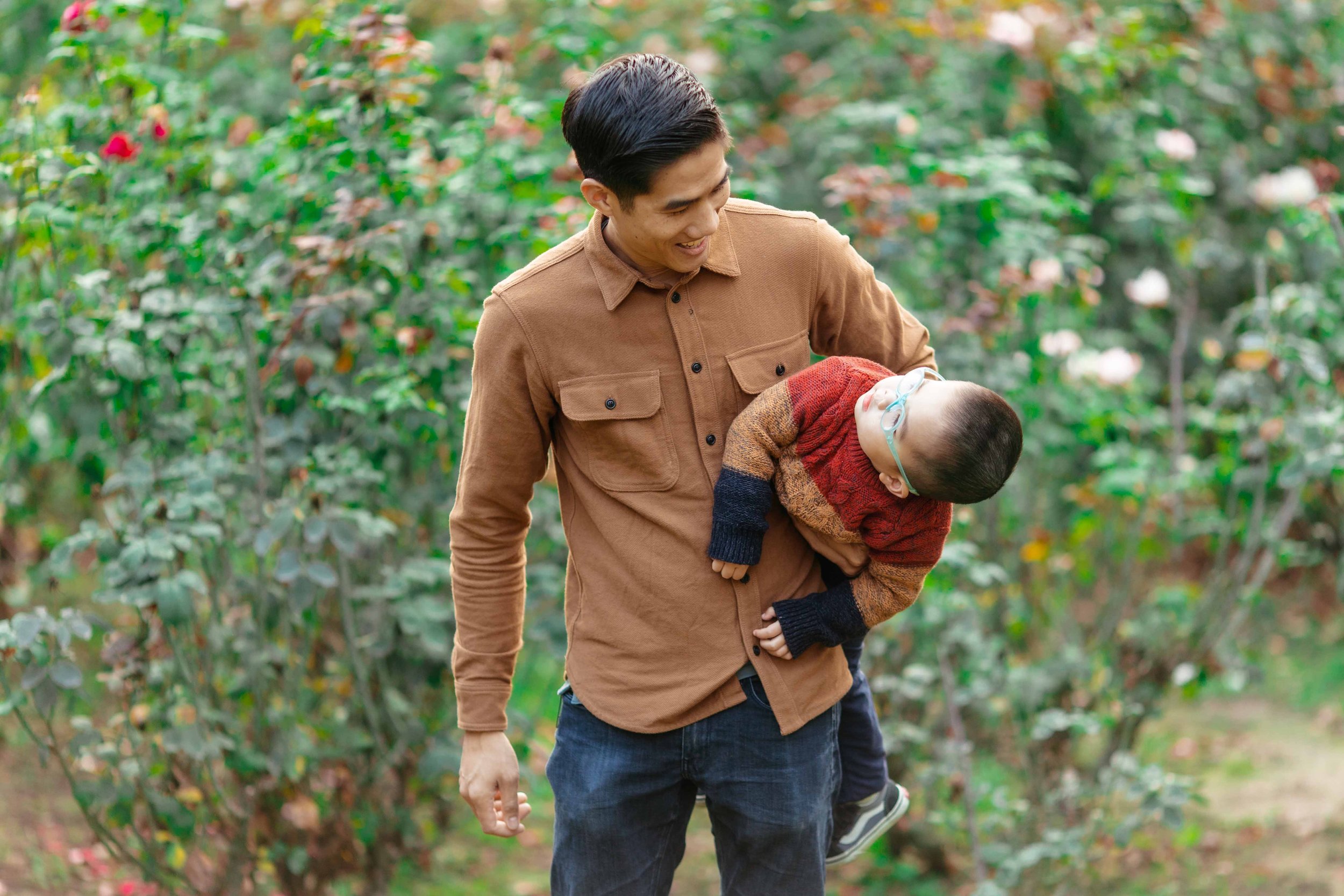A man holding a young boy with glasses, outdoors in a garden or park surrounded by green bushes and flowers.