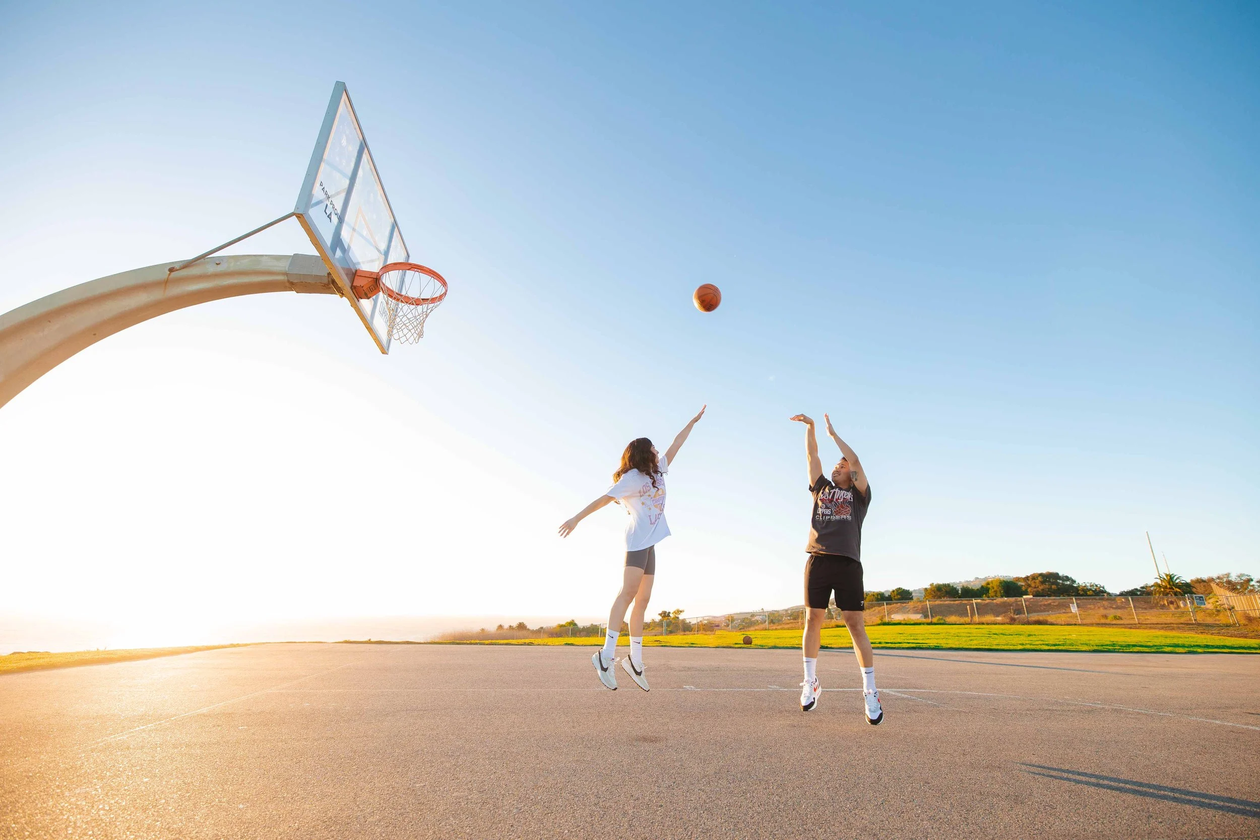 Two people playing basketball on an outdoor court during sunset, with one attempting a shot and the other defending, under a clear blue sky.