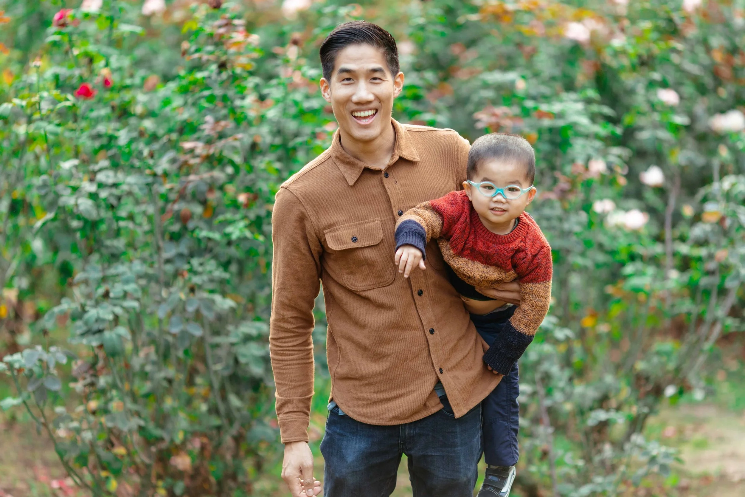 A man with dark hair and a brown shirt smiling while carrying a young boy with glasses, wearing a colorful sweater, through a garden filled with pink and white flowers.