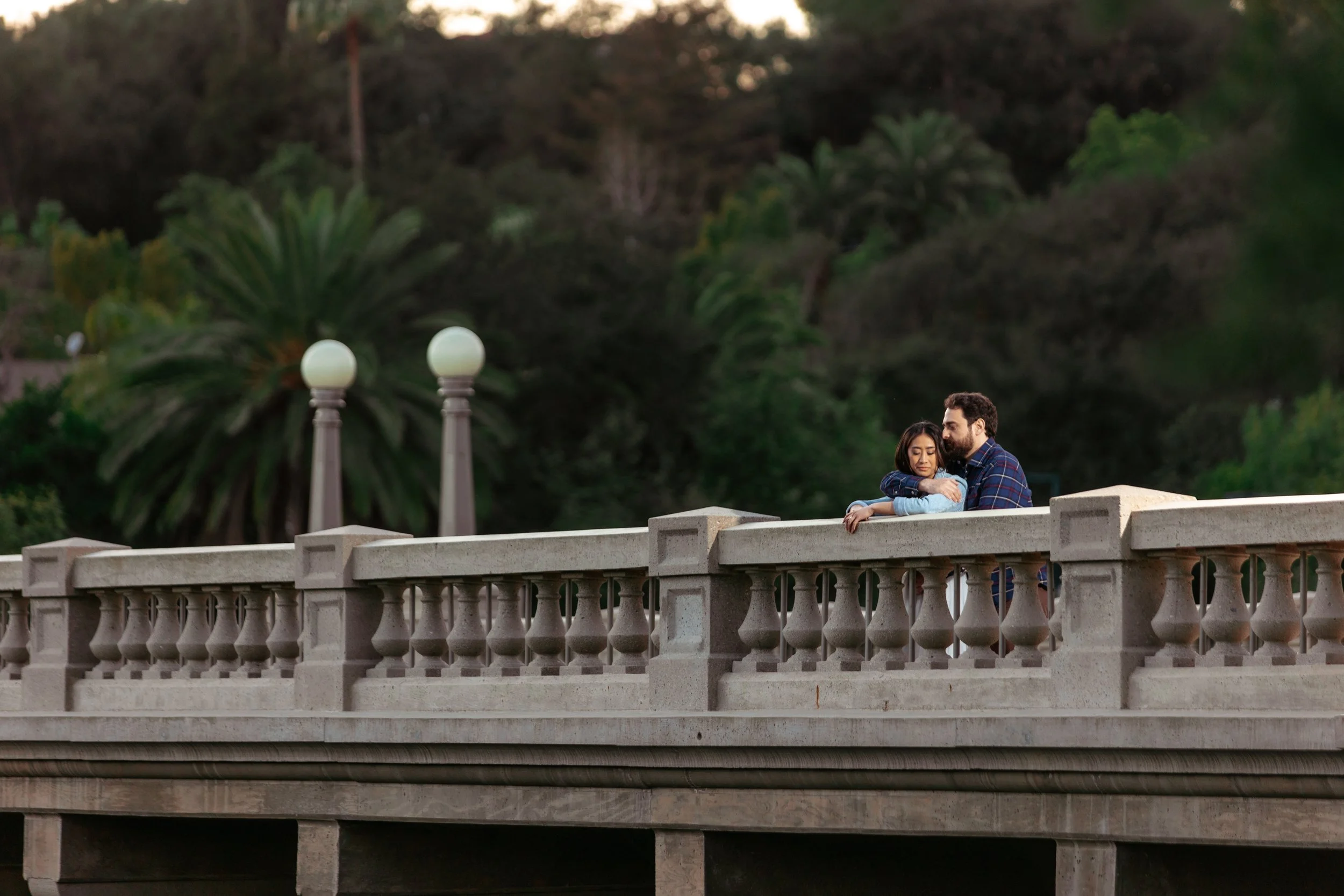 A couple stands on a bridge, embracing each other, with lush green trees and street lamps in the background during dusk.