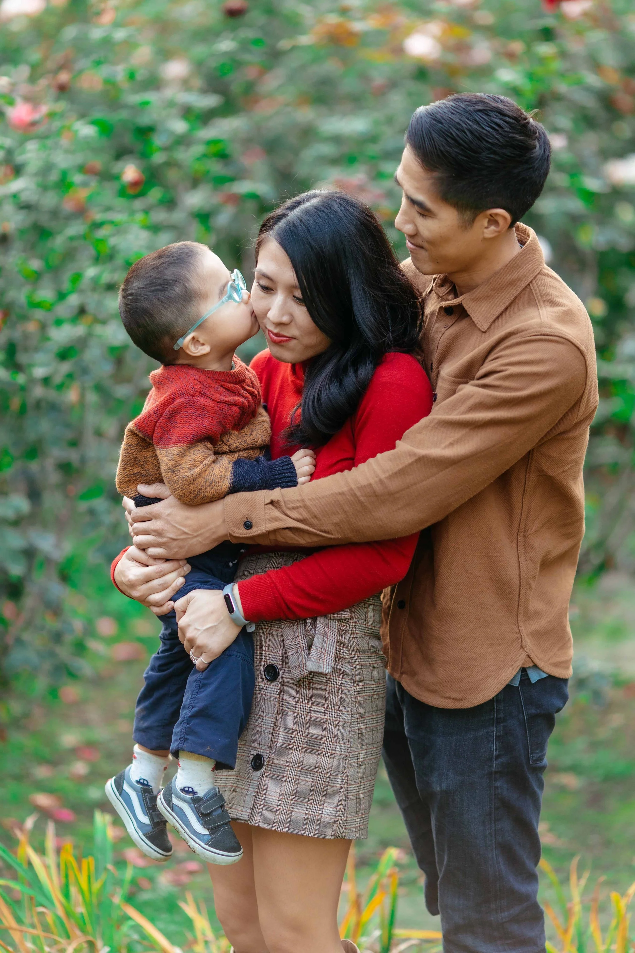 Family of three outdoors; young boy with glasses kissing mother on cheek, father embracing mother, mother smiling, in a garden with green foliage.