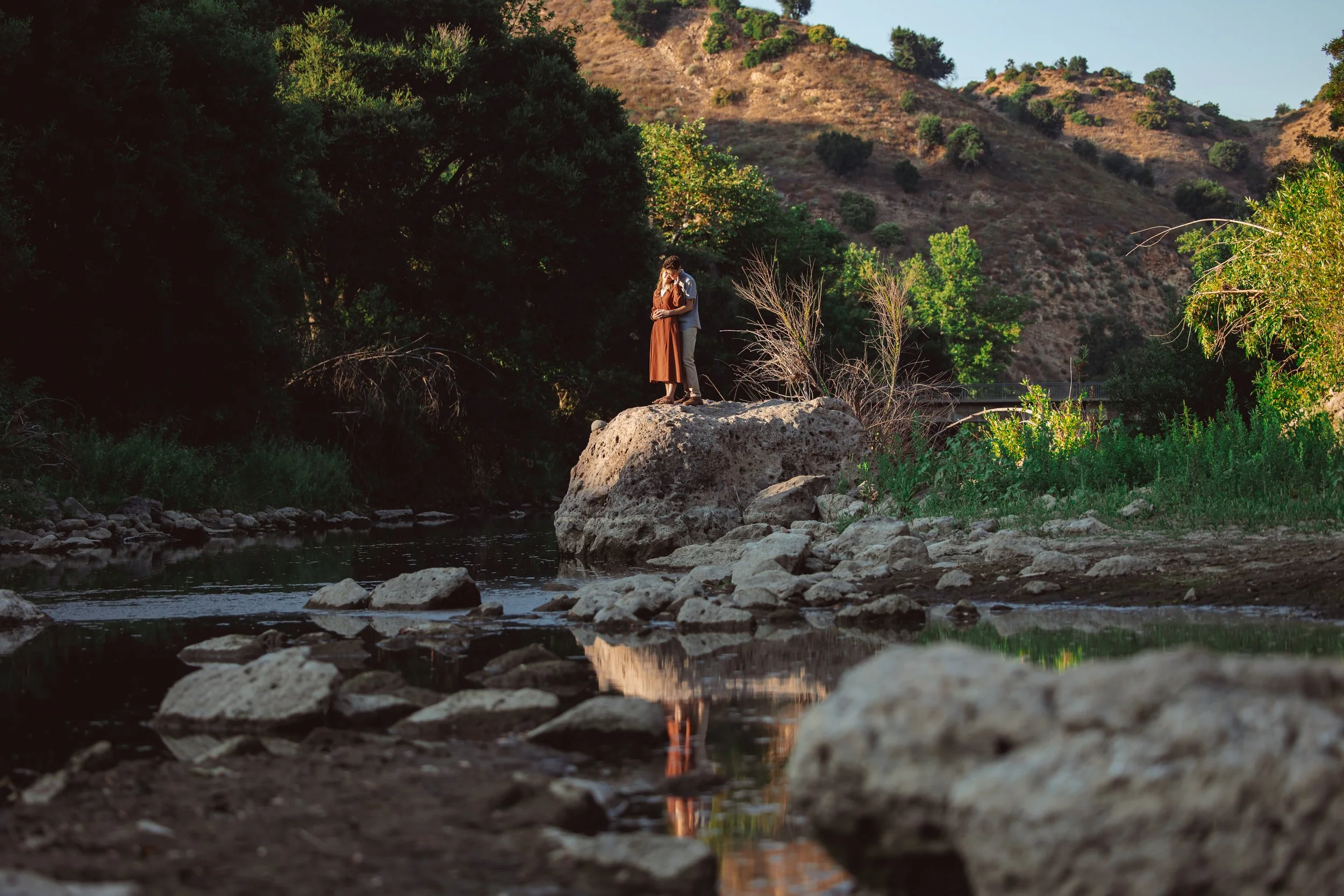 Two people embrace on a large rock in the middle of a river, surrounded by trees and hills, with the river reflecting their image.