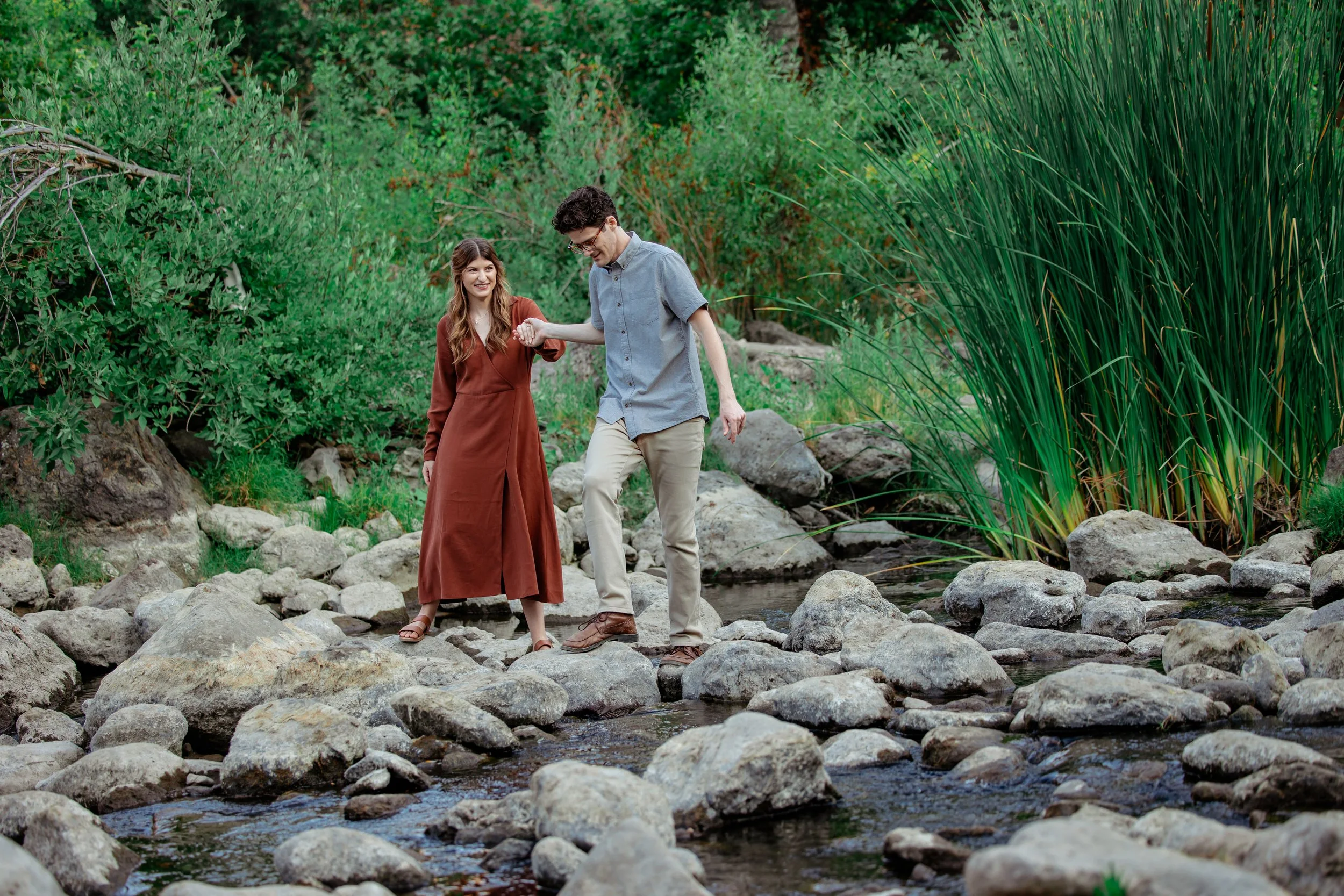 A man and woman walking on rocks across a small creek in a natural, green outdoor setting, with the man helping the woman balance as they walk.
