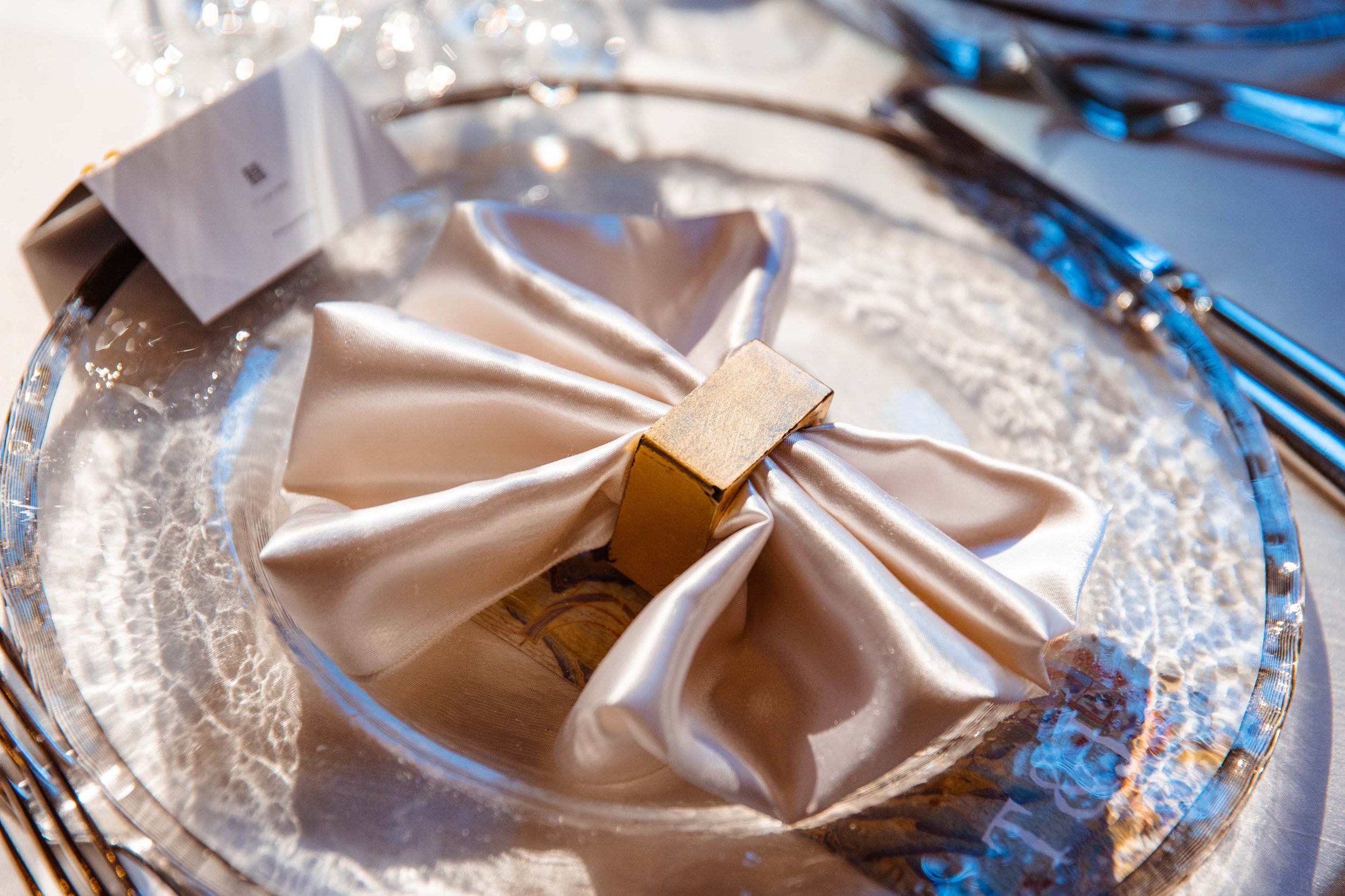 A silver charger plate with a wrapped napkin tied with a golden square ring, set on a table with silverware.