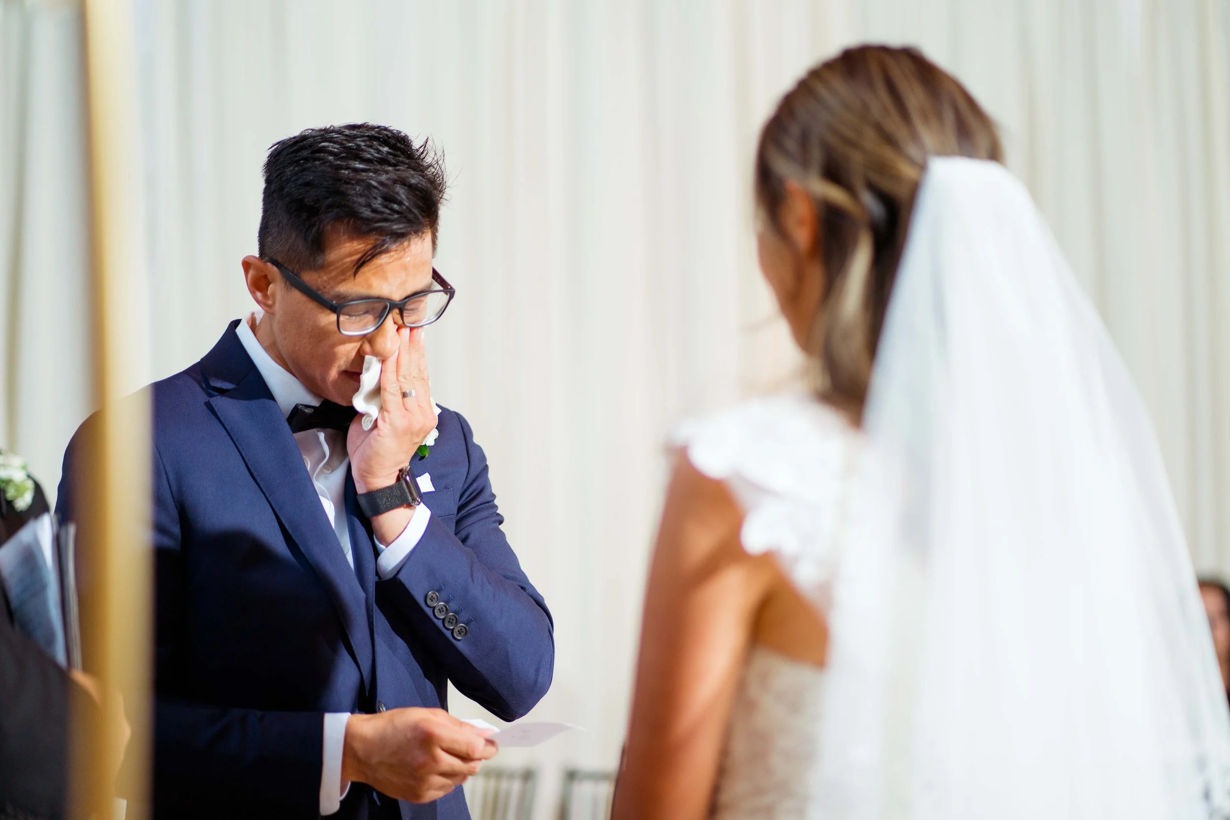 A groom in a navy suit is emotional at a wedding, wiping tears with a tissue while holding a piece of paper; the bride in a white dress and veil is partially visible.