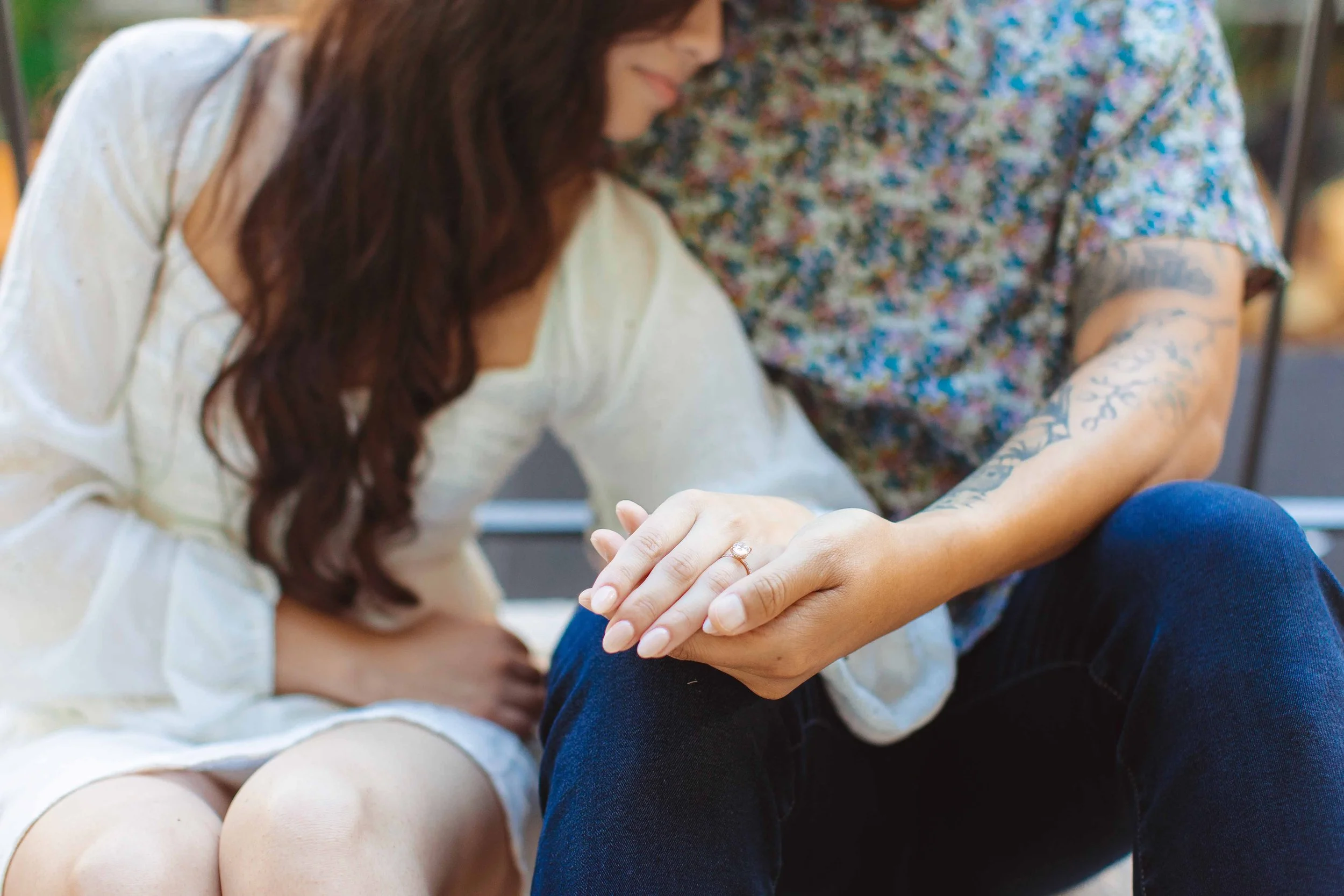 A woman with long, wavy brown hair and tattoos on her arm holding hands with another person. The woman is wearing a white dress, and the second person is wearing a floral shirt and dark jeans, with a ring on their finger.