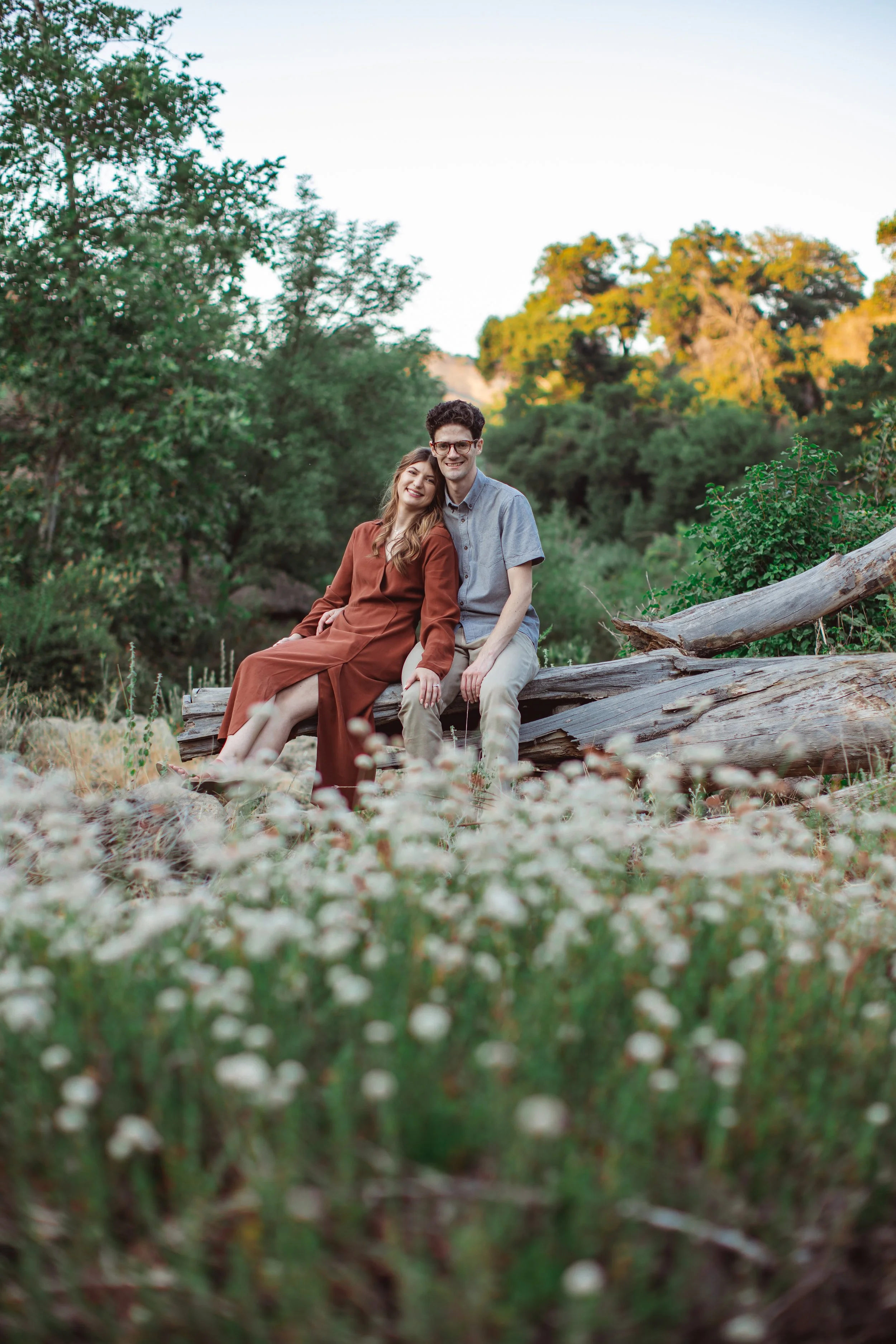 A happy couple sitting on a fallen log in a lush, green outdoor setting at dusk, surrounded by trees and white wildflowers.