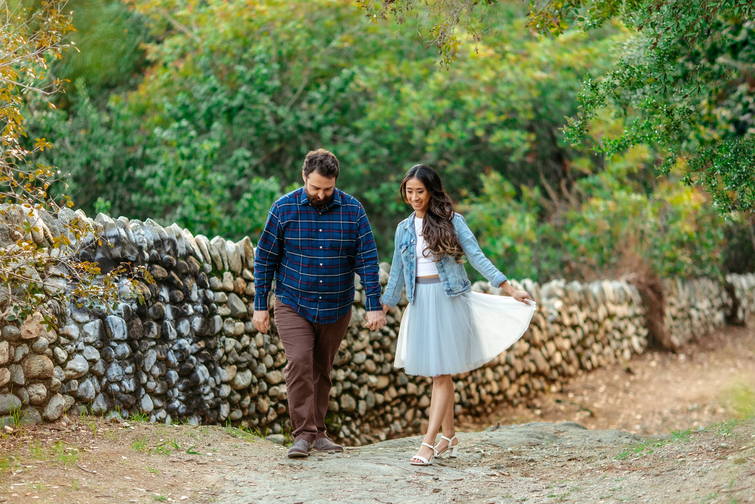 A man and woman walking hand in hand on a dirt path beside a stone wall, surrounded by green trees and foliage.