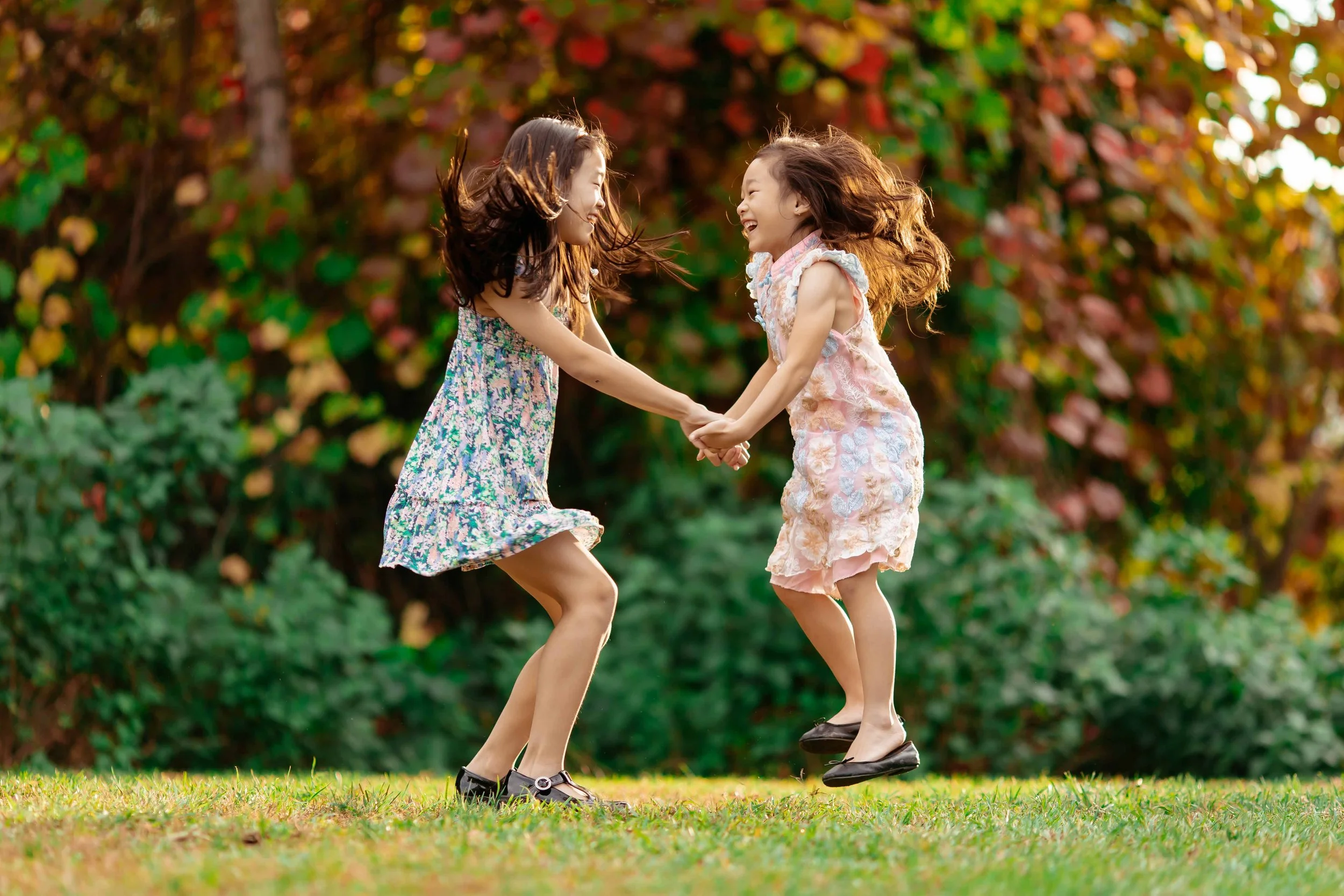 Two young girls in floral dresses playing and holding hands in a park with green grass and colorful autumn trees.