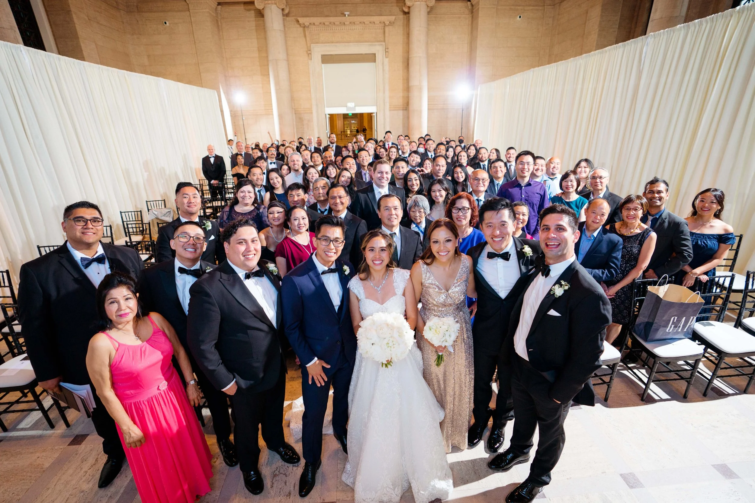 A large wedding group photo featuring bride and groom in the front center, surrounded by friends and family banquet style in a grand hall with cream curtains.