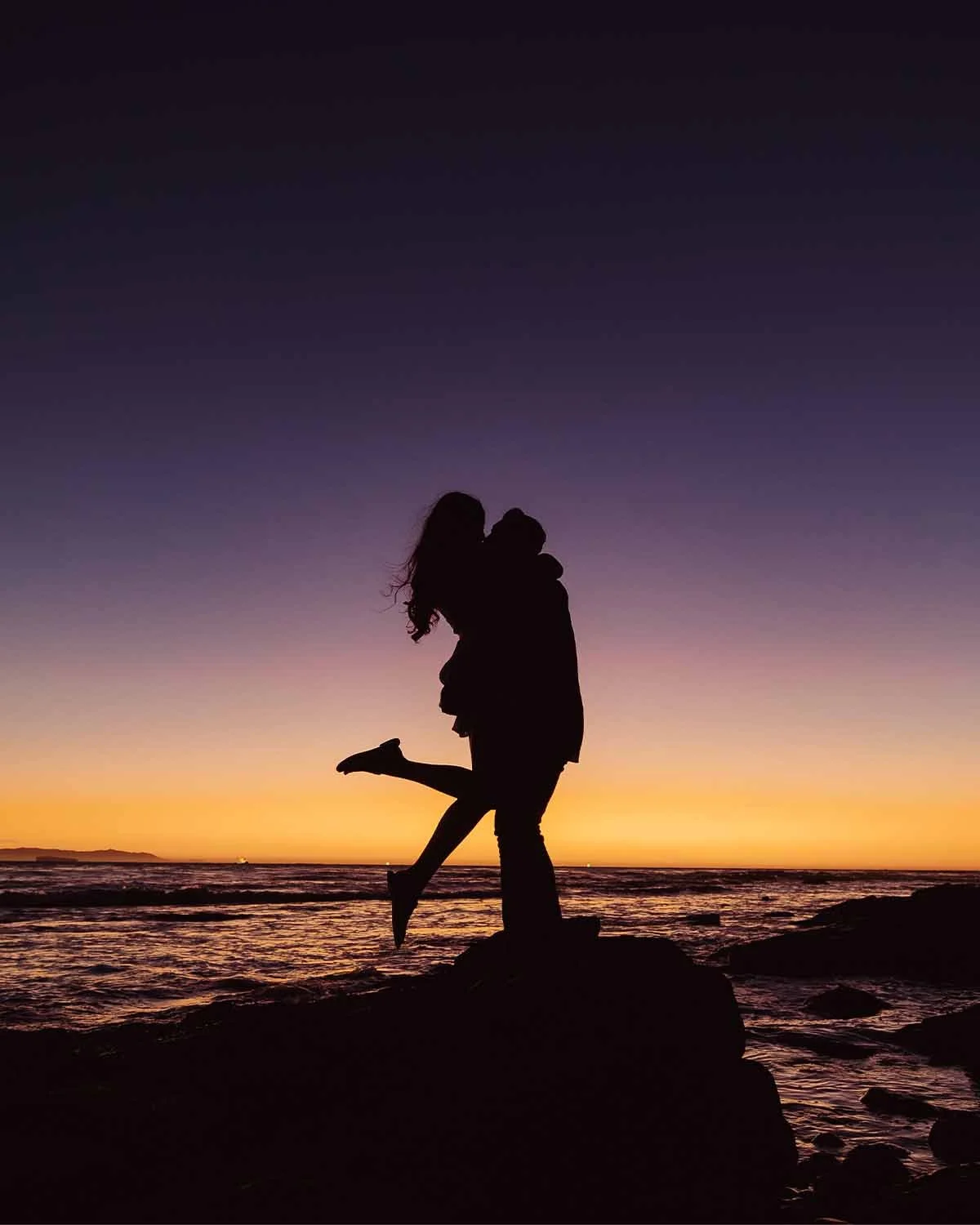 Silhouette of a couple embracing on rocks by the beach during sunset, with purple, orange, and pink sky