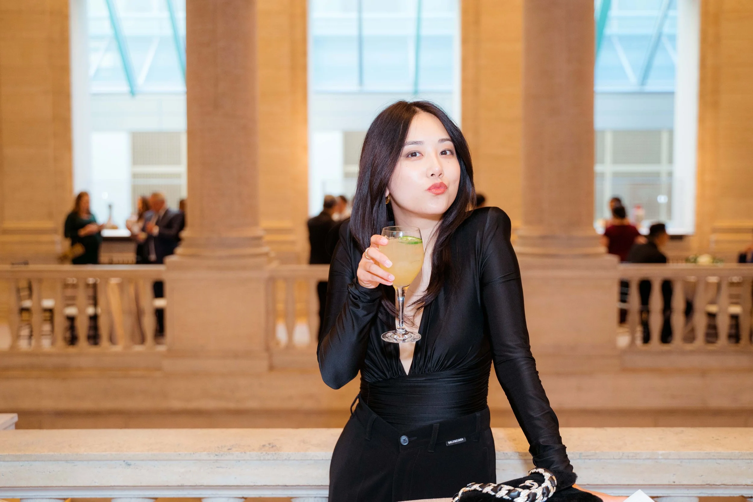 A woman with black hair in a black dress holding a cocktail in a upscale setting with large columns and other people in the background.