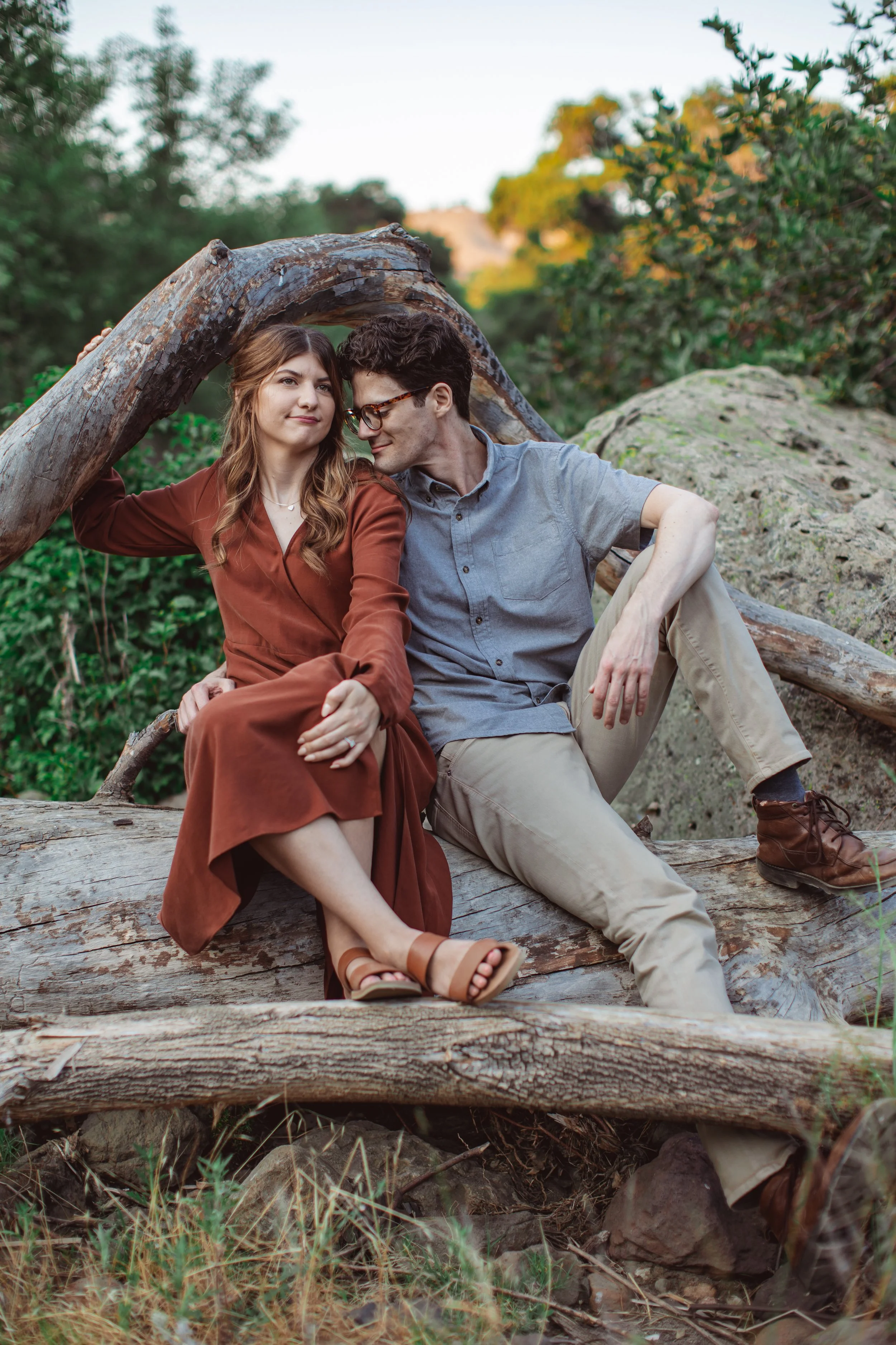 A young woman in a brown dress and tan sandals sitting on a log next to a young man in a gray shirt and beige pants, with trees and rocks in the background.