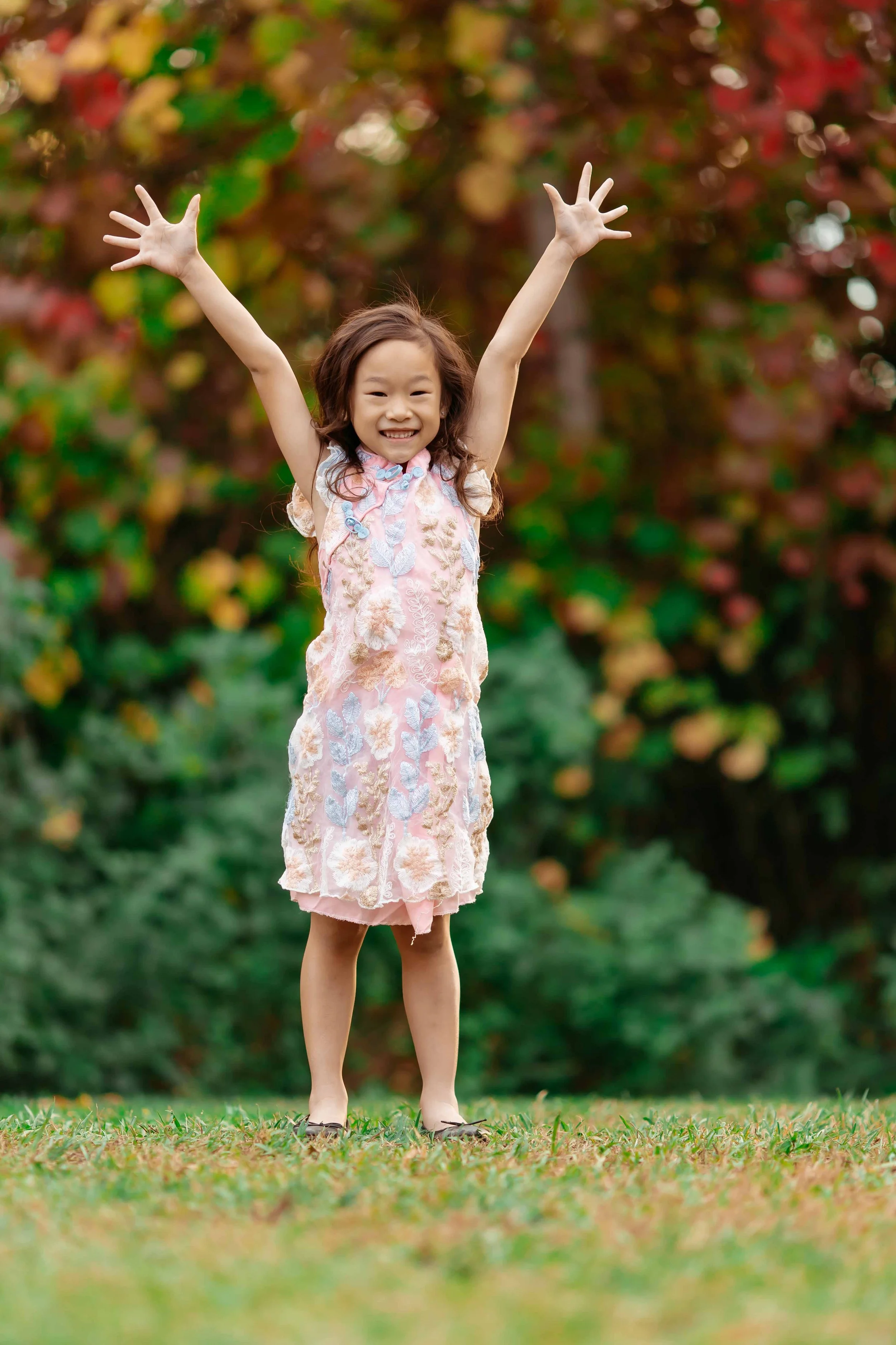 A young girl with long hair smiling and raising her arms in a field of grass, with trees in autumn colors in the background.