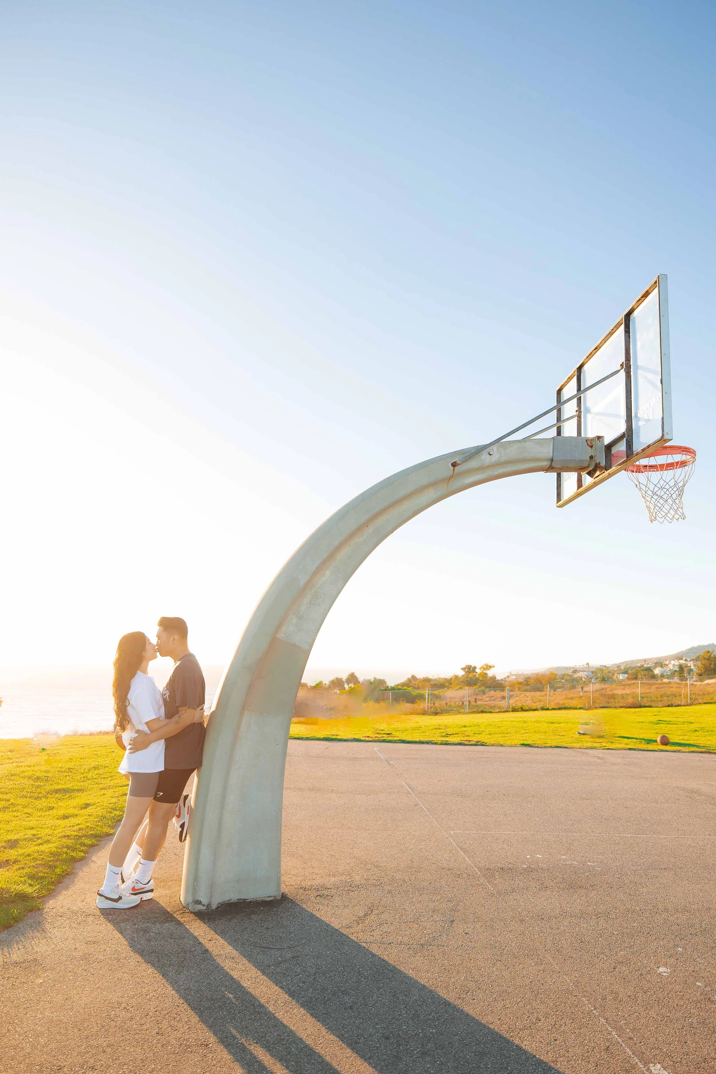 A couple kissing on an outdoor basketball court at sunset, with a basketball hoop and a scenic landscape in the background.