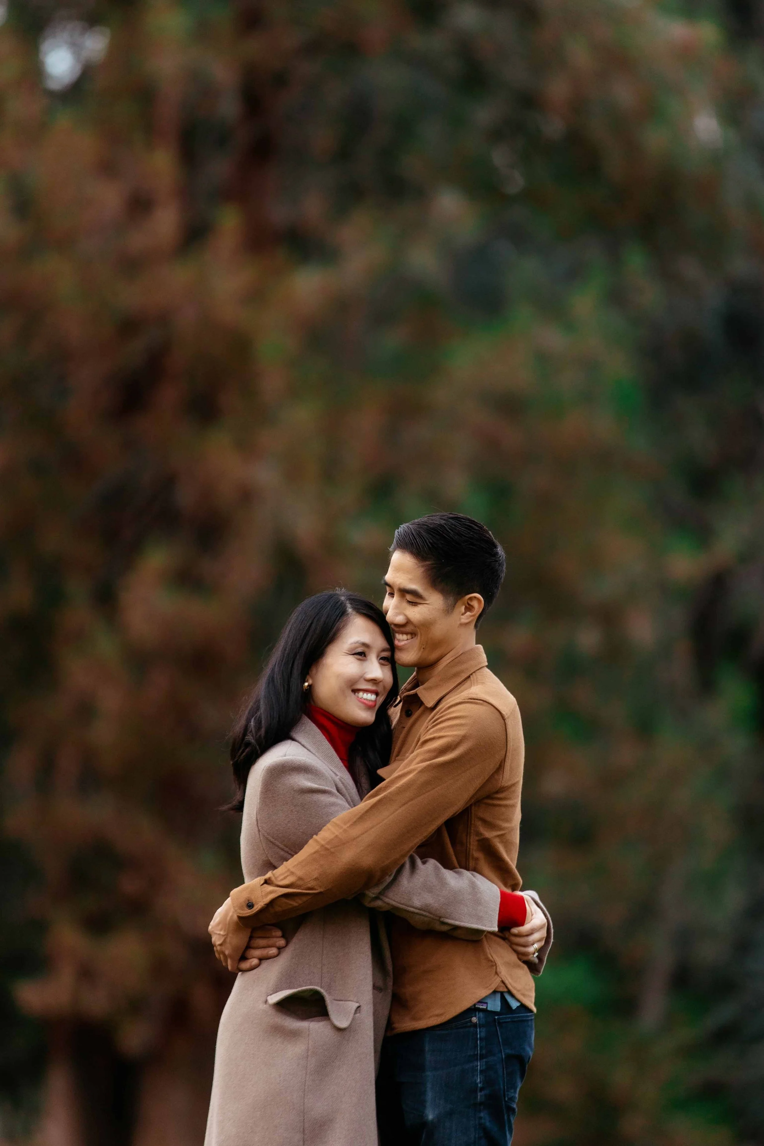 A smiling couple hugging outdoors during autumn, with trees and colorful fall foliage in the background.