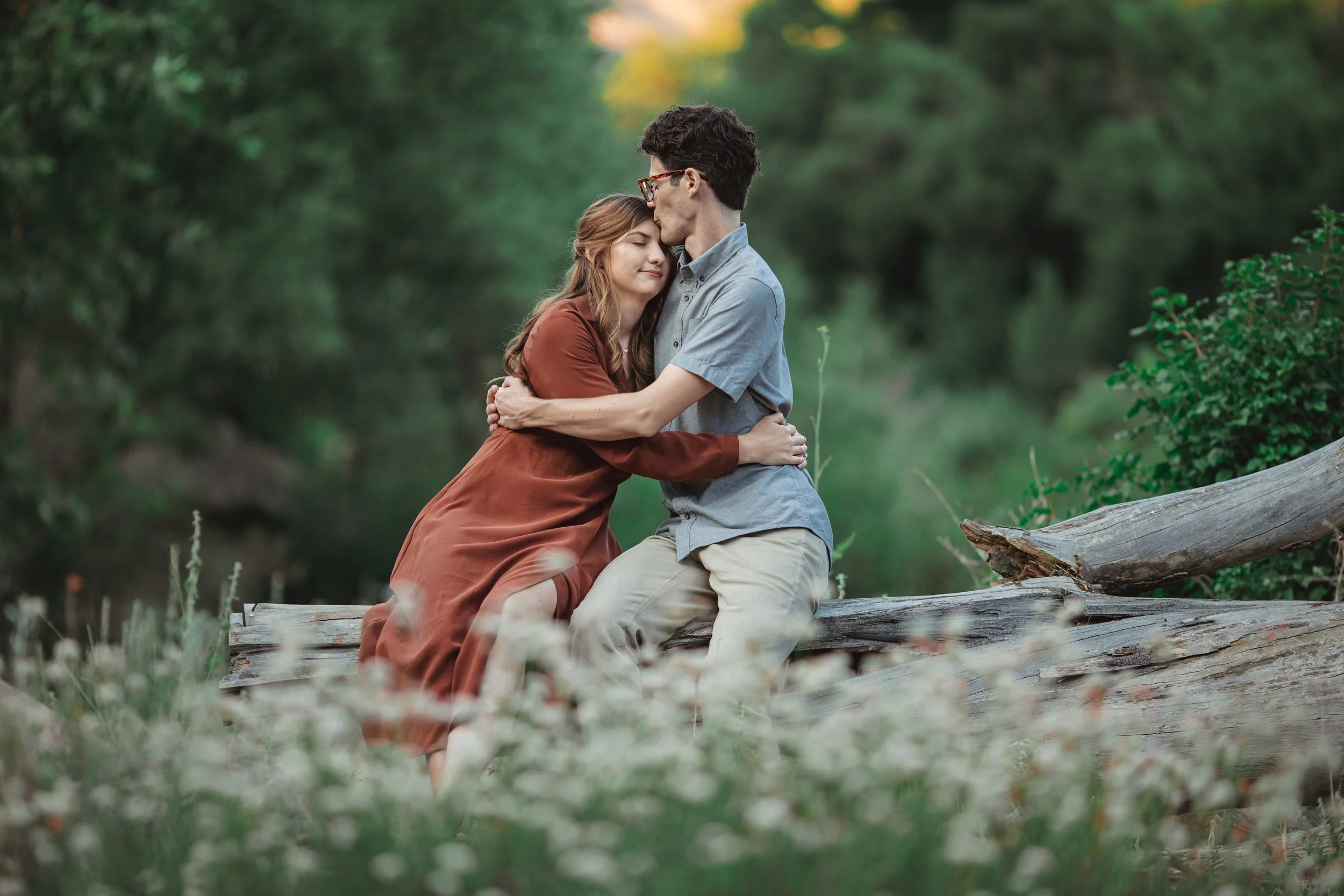 A couple hugging on a fallen log in a lush green forest, with the man kissing the woman's forehead and the woman smiling gently, dressed in casual clothing.