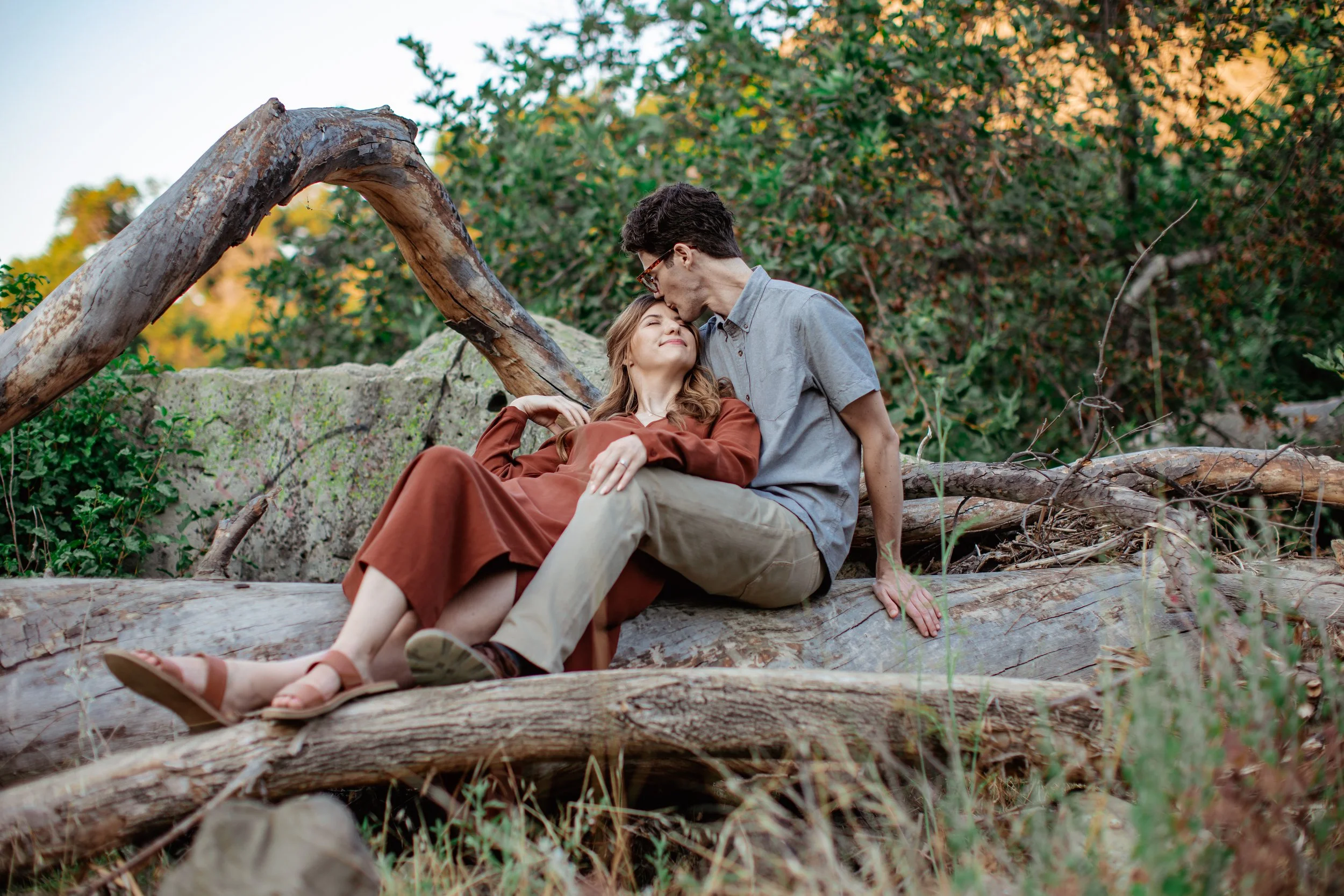 A young couple sitting on a fallen tree trunk in a forested area, with the man tenderly kissing the woman's forehead as she leans into him. The woman wears a rust colored dress and sandals, and the man is dressed in a light gray shirt and beige pants
