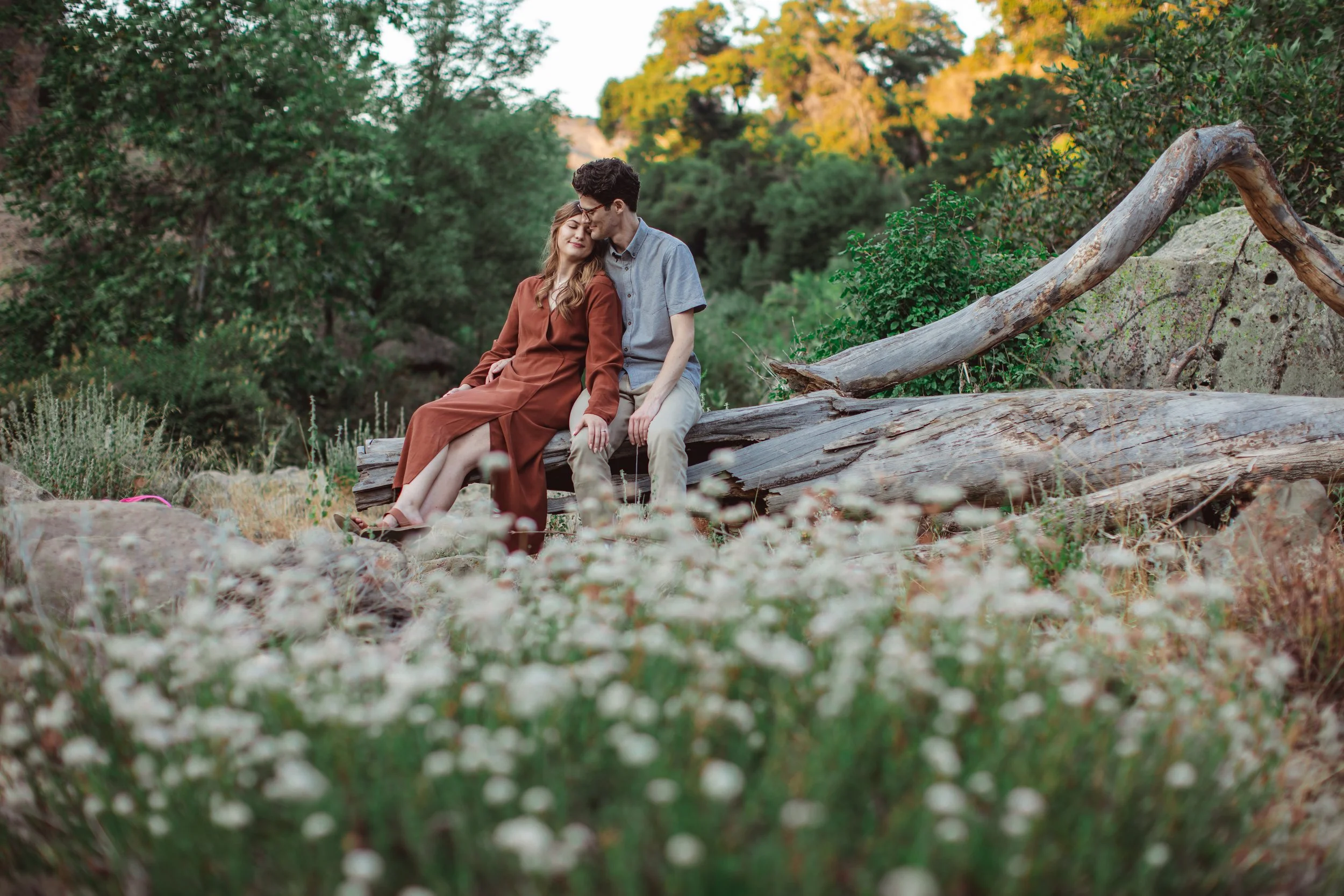 A young couple sitting closely together on a fallen log in a natural outdoor setting surrounded by greenery and wildflowers.