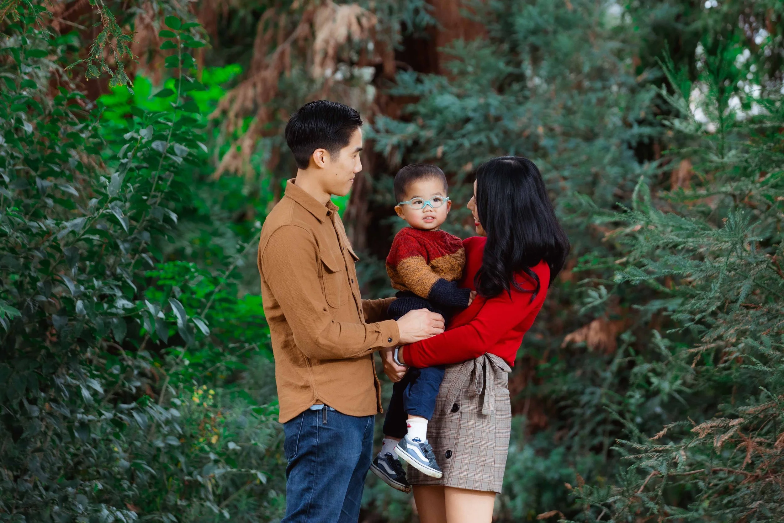 A family of three stands in a lush green forest. The father and mother hold their young son, who is wearing glasses, a red and orange sweater, and dark pants. The mother is dressed in a red top and plaid skirt. They are enjoying a moment together out