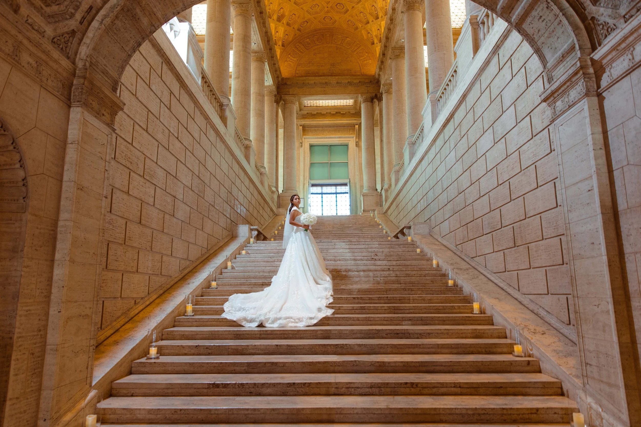 A bride in a white wedding gown holding a bouquet standing on a grand marble staircase inside a historic building with high ceilings and large windows.