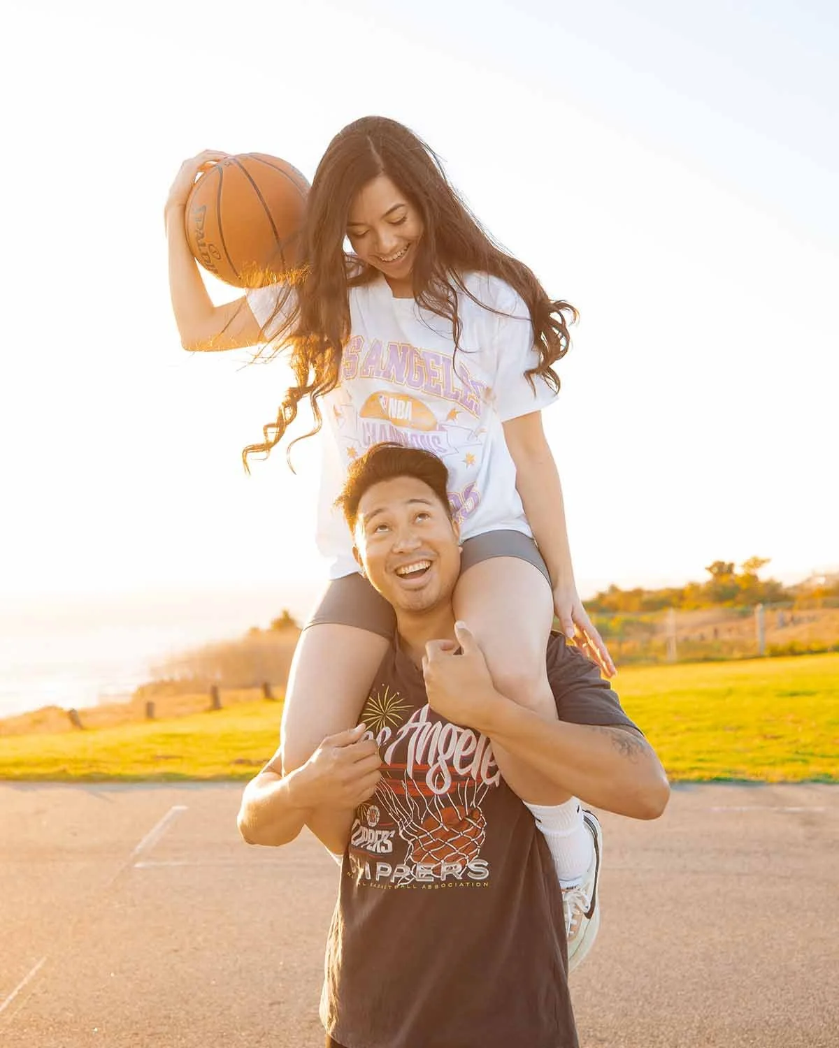 A man carrying a woman on his shoulders during sunset on a basketball court, with both smiling and looking at each other.