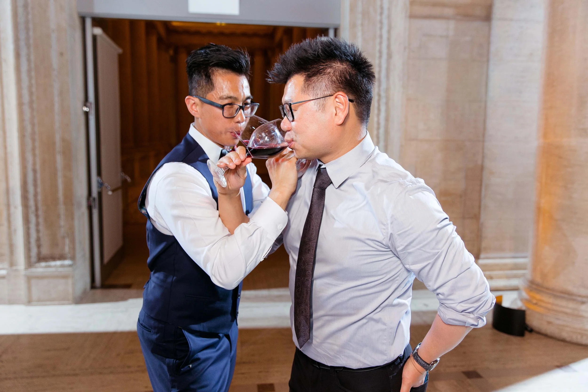 Two men, one in a tuxedo and the other in a shirt and tie, drinking red wine together in an elegant setting.