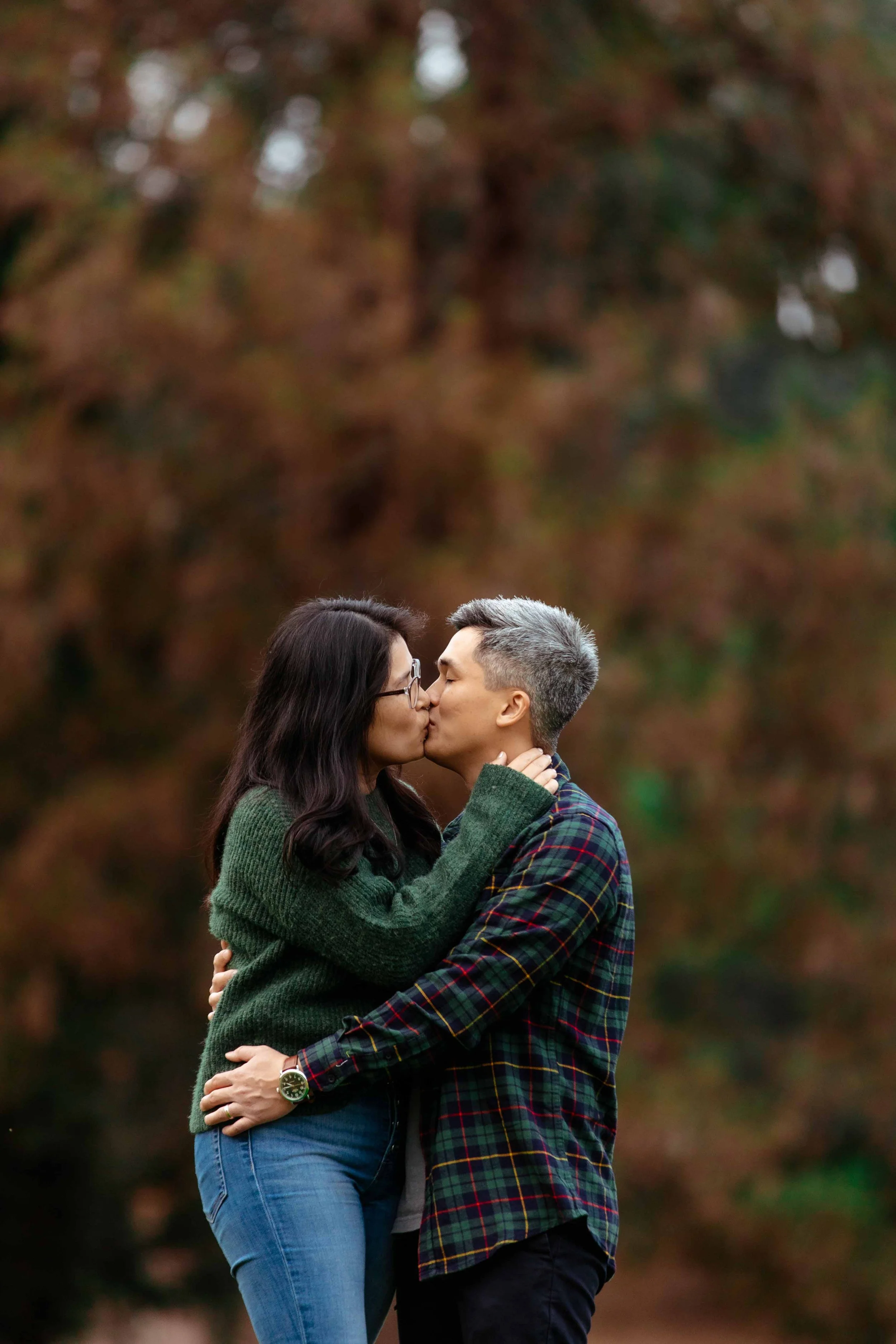 A couple kissing outdoors with autumn trees in the background.