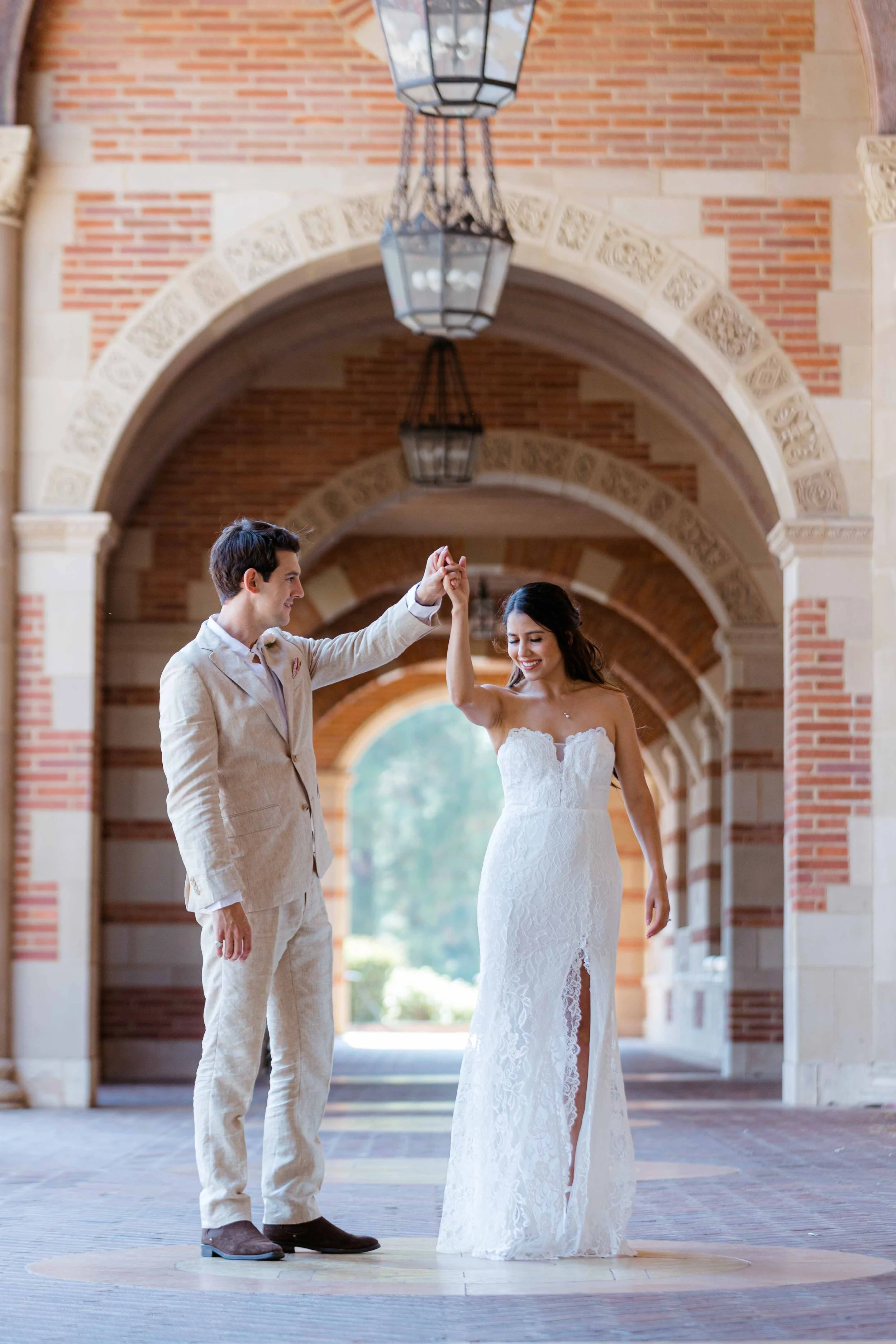 A bride and groom dancing under brick arches, with the groom in a light suit and the bride in a strapless white lace wedding dress.