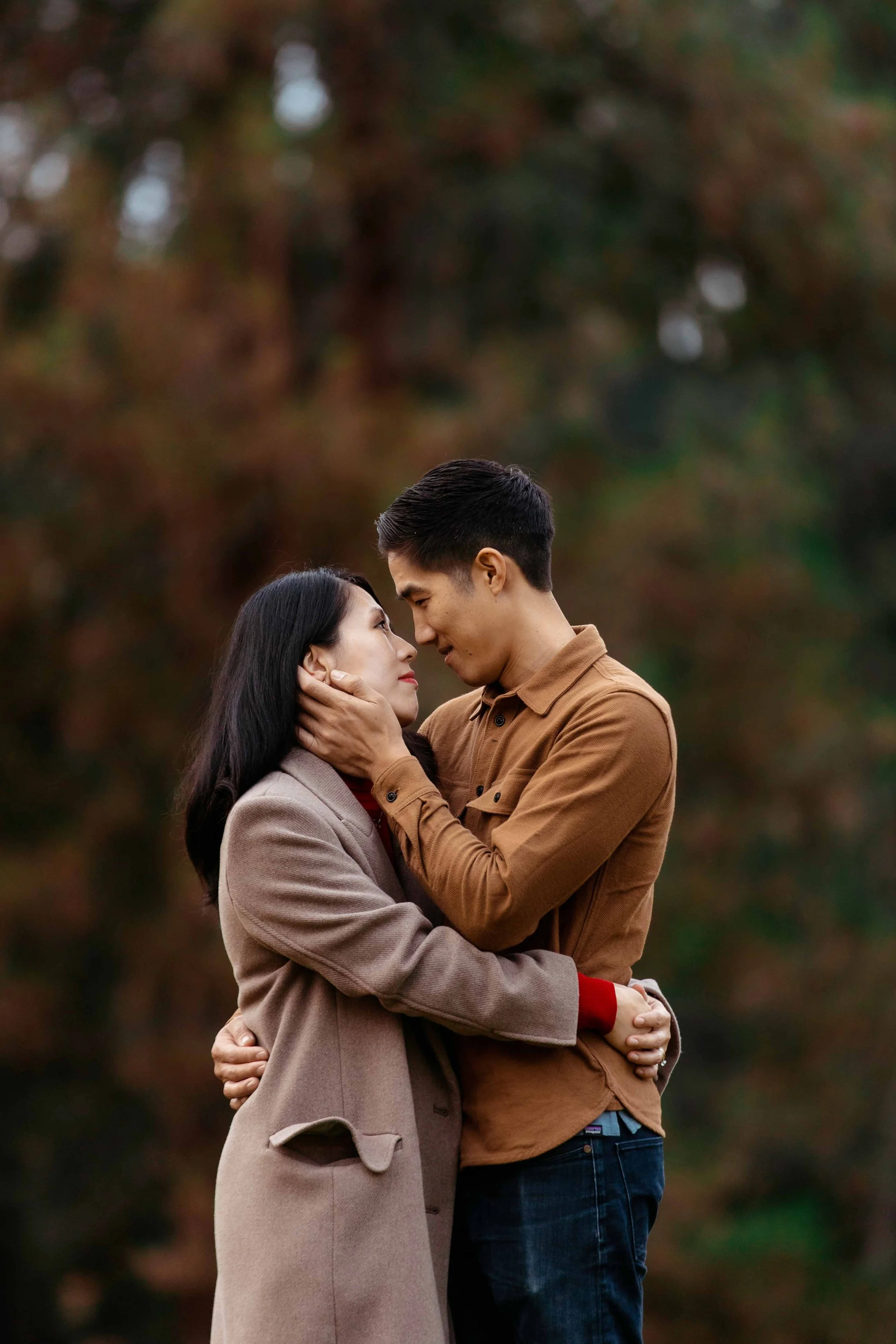 A man and woman are embracing outdoors, gazing into each other's eyes in a close, intimate moment during autumn.