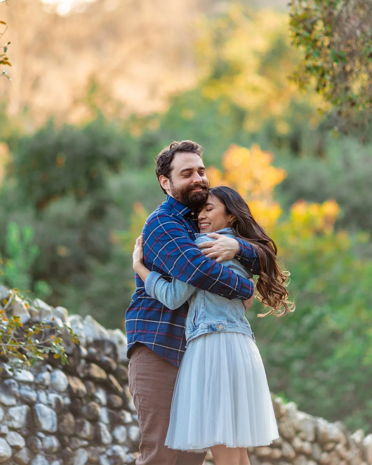 A man and woman hugging outdoors on a fall day with trees and a stone wall in the background.